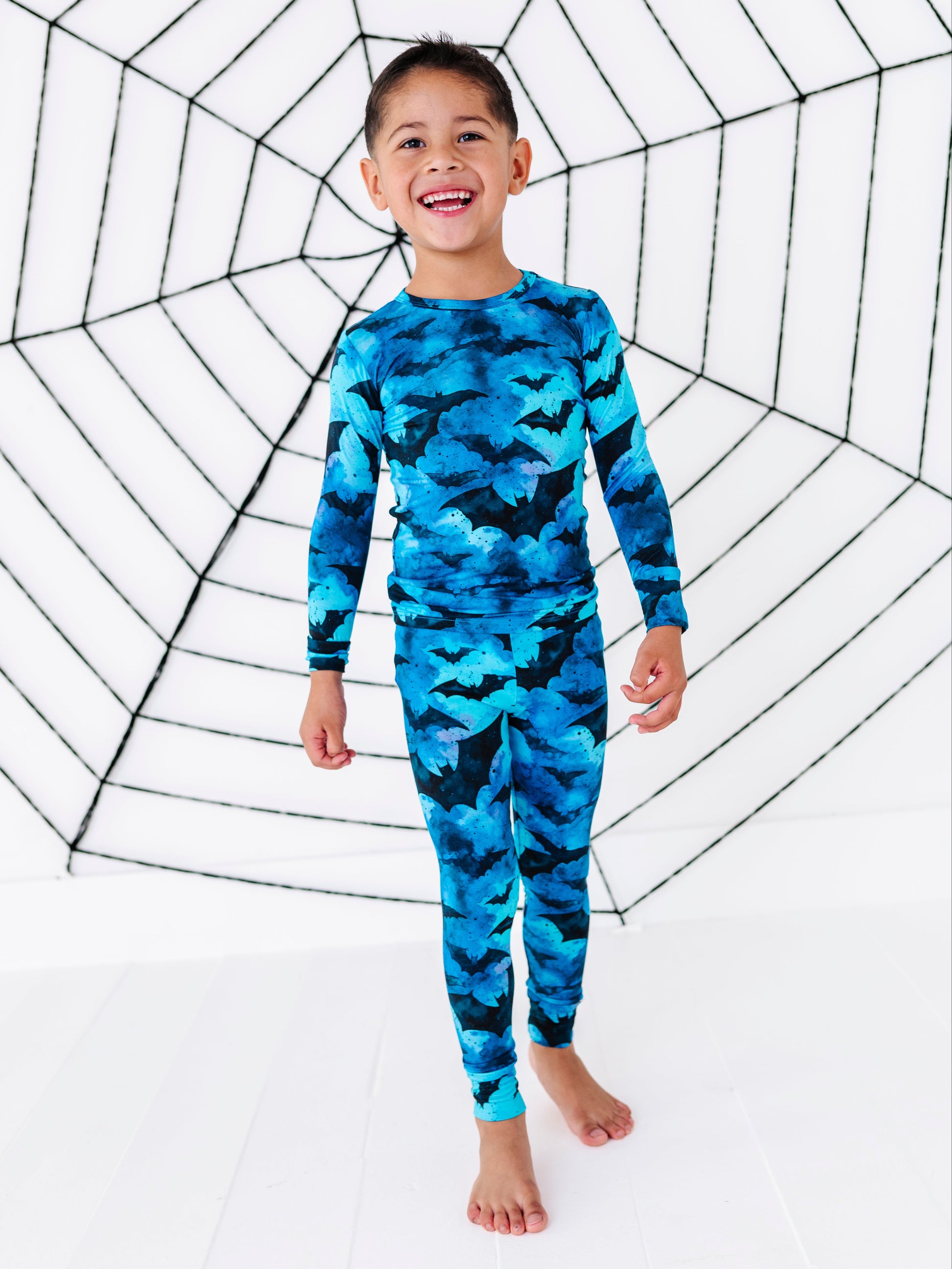 A young boy smiling and wearing a blue and black bat-patterned long-sleeve pajama set, standing barefoot in front of a large black spiderweb graphic on a white background.