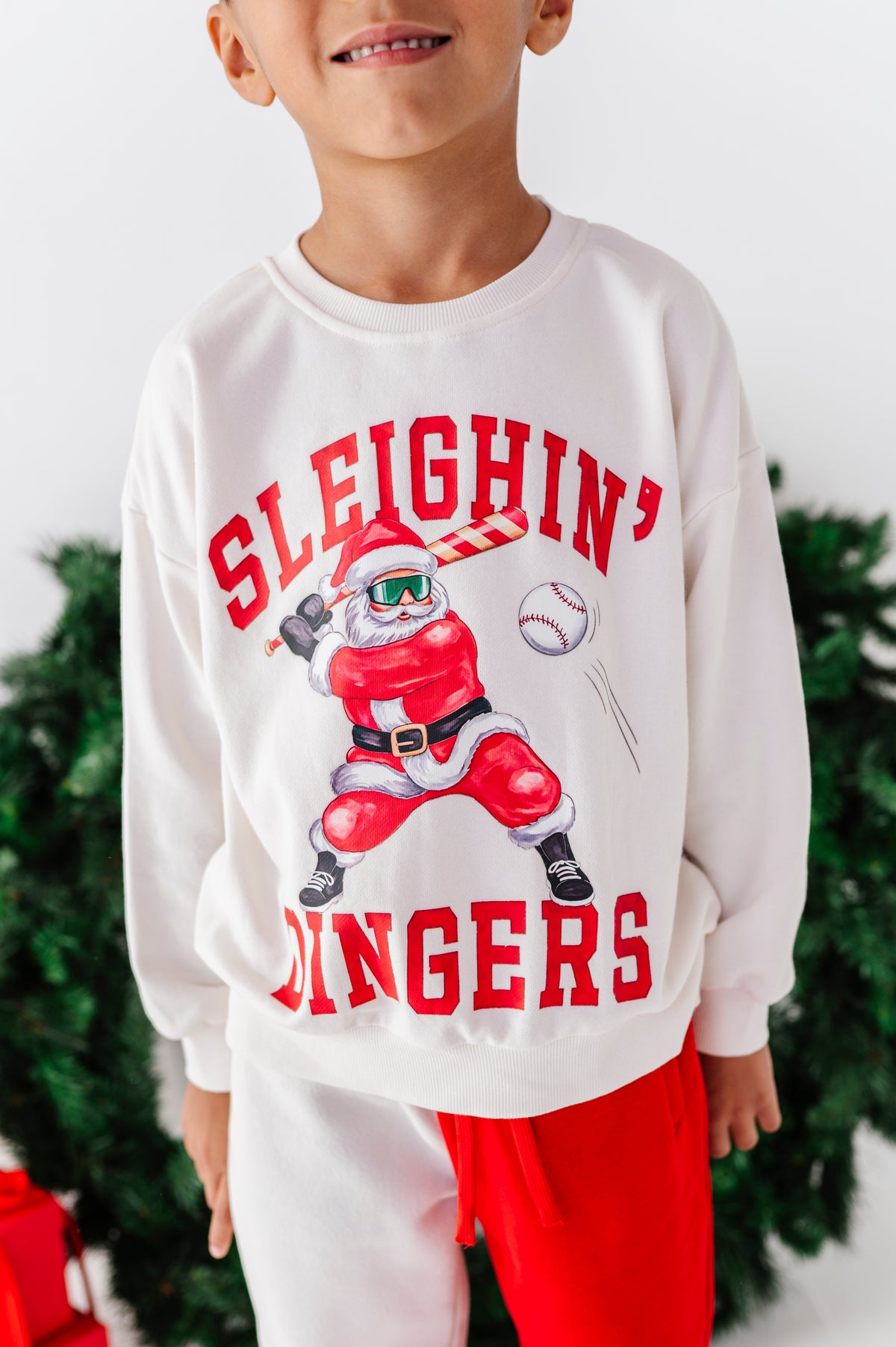 A child wearing a white crewneck sweatshirt with a red graphic of a baseball player dressed as Santa and the text 'SLEIGHIN' BINGERS', paired with red pants, with a blurred Christmas tree in the background.