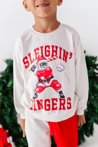 A child wearing a white crewneck sweatshirt with a red graphic of a baseball player dressed as Santa and the text 'SLEIGHIN' BINGERS', paired with red pants, with a blurred Christmas tree in the background.