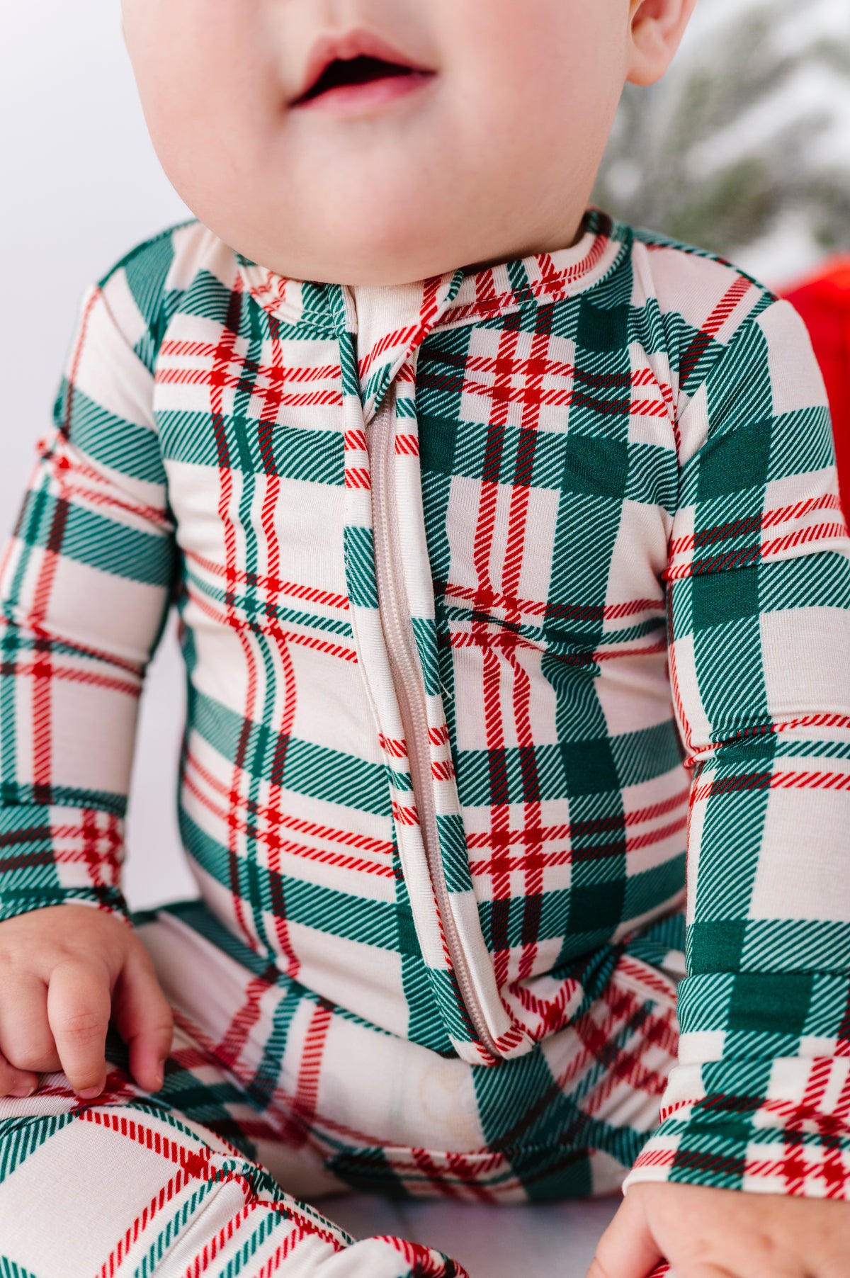 A close-up of a baby wearing a festive zip-up bamboo sleeper with a green, red, and white plaid pattern.