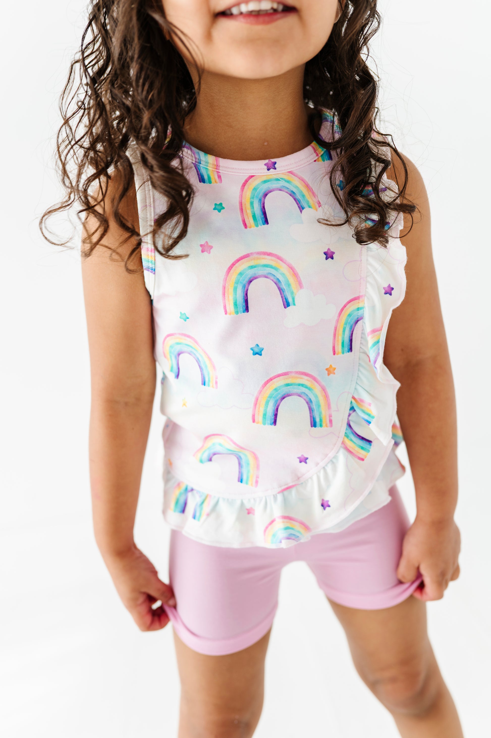 Little girl wearing a sleeveless peplum top with watercolor rainbow patterns and pink bamboo biker shorts on a white background