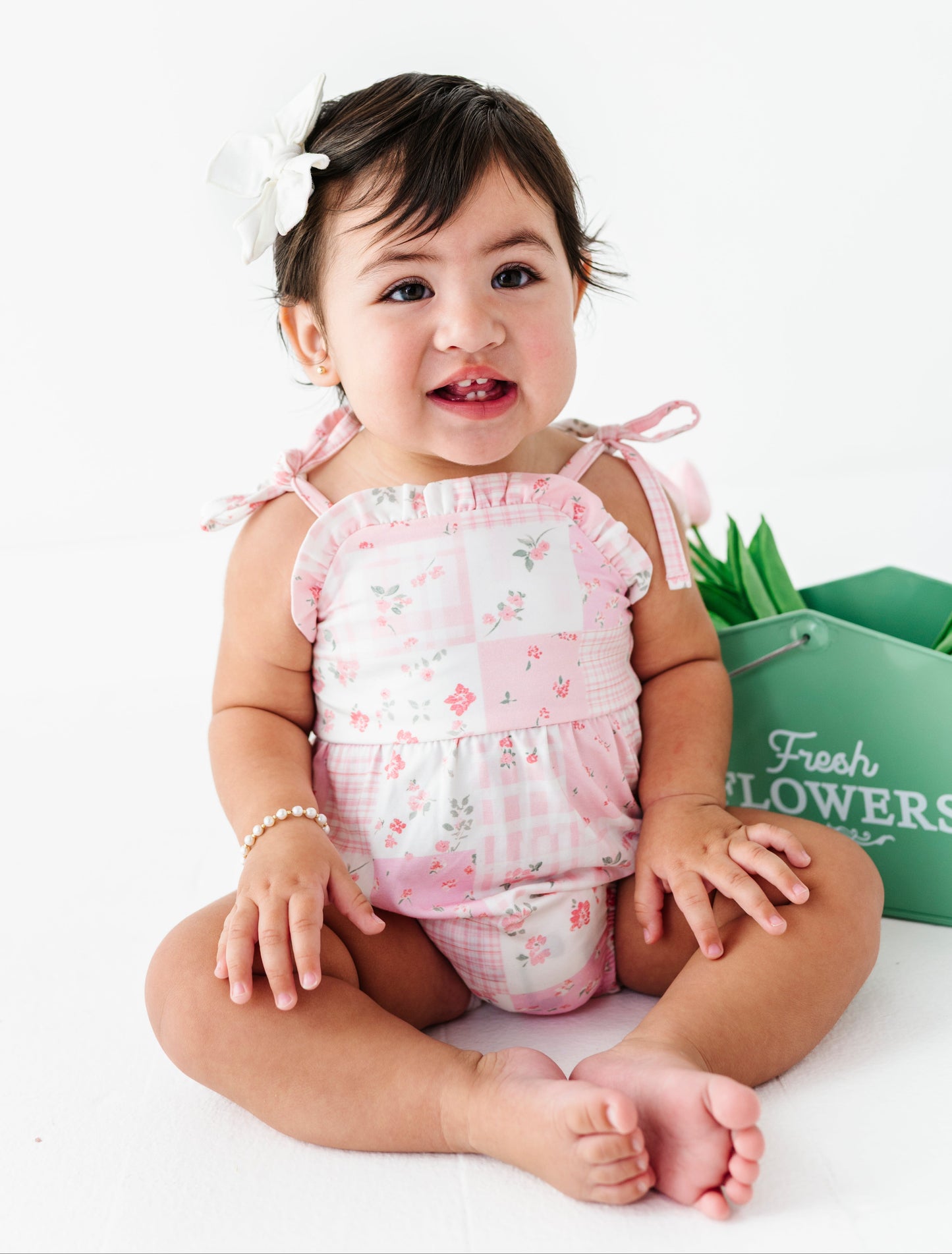 Baby girl in a pink floral bamboo romper sitting on a white surface with a green bucket of flowers in the background.