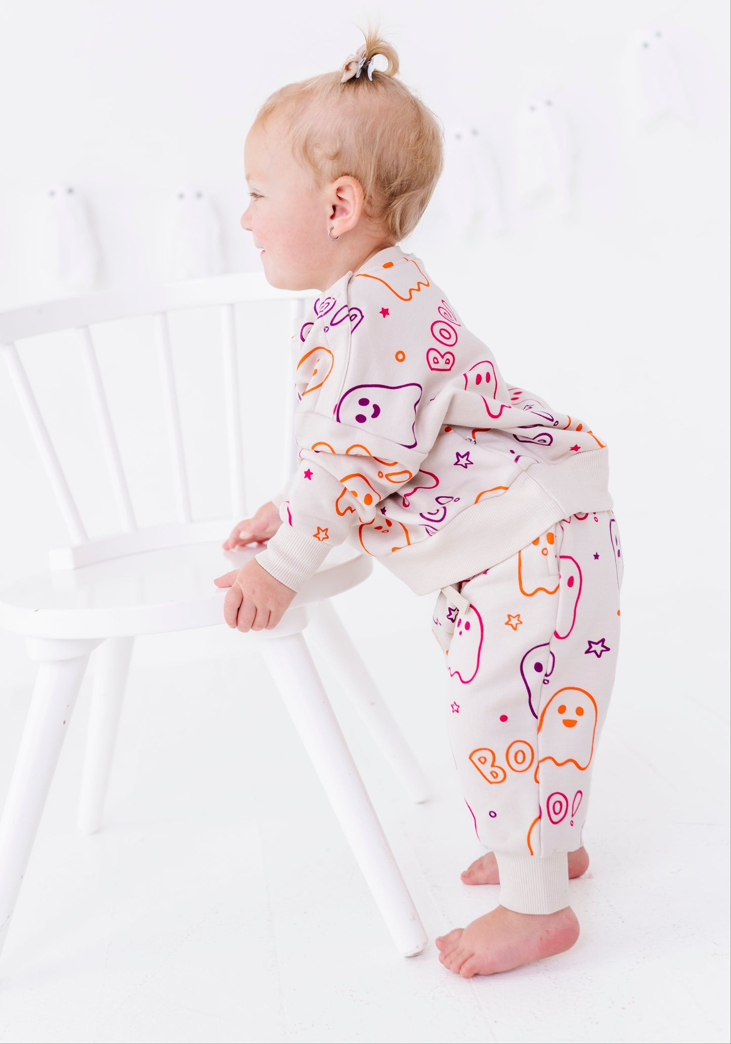 A toddler standing barefoot next to a white chair, wearing a cream-colored long-sleeve sweatshirt and matching pants with a colorful ghost and 'BOO' Halloween pattern.