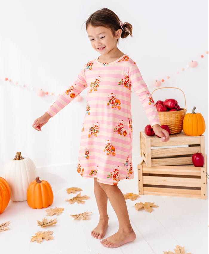 A young girl stands barefoot in a studio setting wearing a pink long-sleeve pajama dress with a gingham check pattern and a print of cartoon turkeys. The background is decorated with autumn elements including pumpkins, fall leaves, a wooden crate with red apples, and a pink balloon garland.