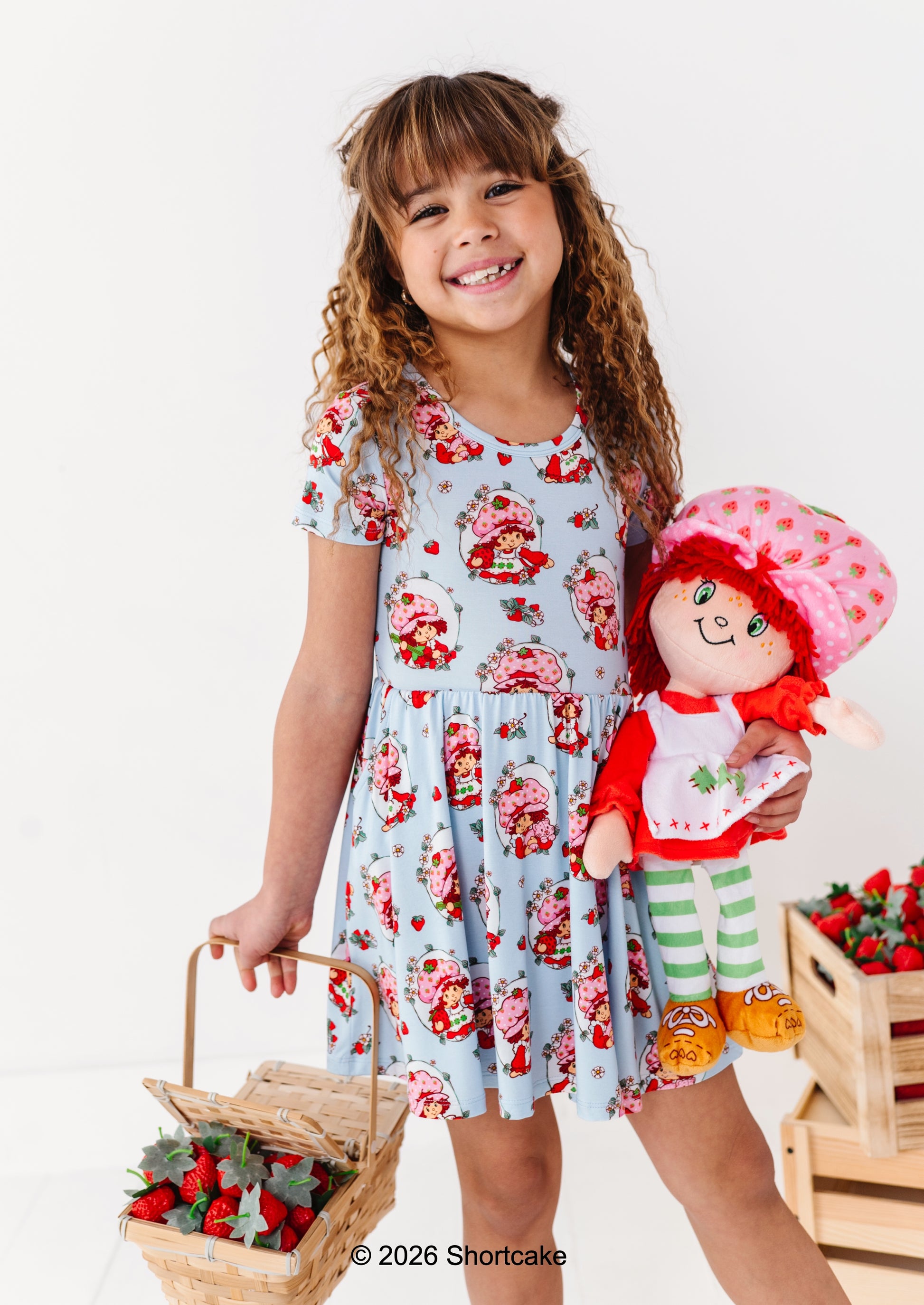 Young girl in a licensed Strawberry Shortcake™ bamboo dress holding a Strawberry Shortcake™ doll and basket of strawberries on a white background