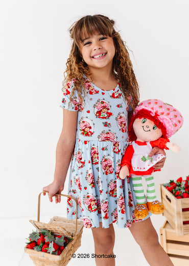 Young girl in a licensed Strawberry Shortcake™ bamboo dress holding a Strawberry Shortcake™ doll and basket of strawberries on a white background