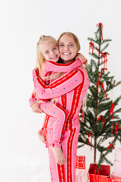 A woman and a young girl wearing matching pink and red striped bamboo holiday pajamas hugging in front of a decorated Christmas tree.
