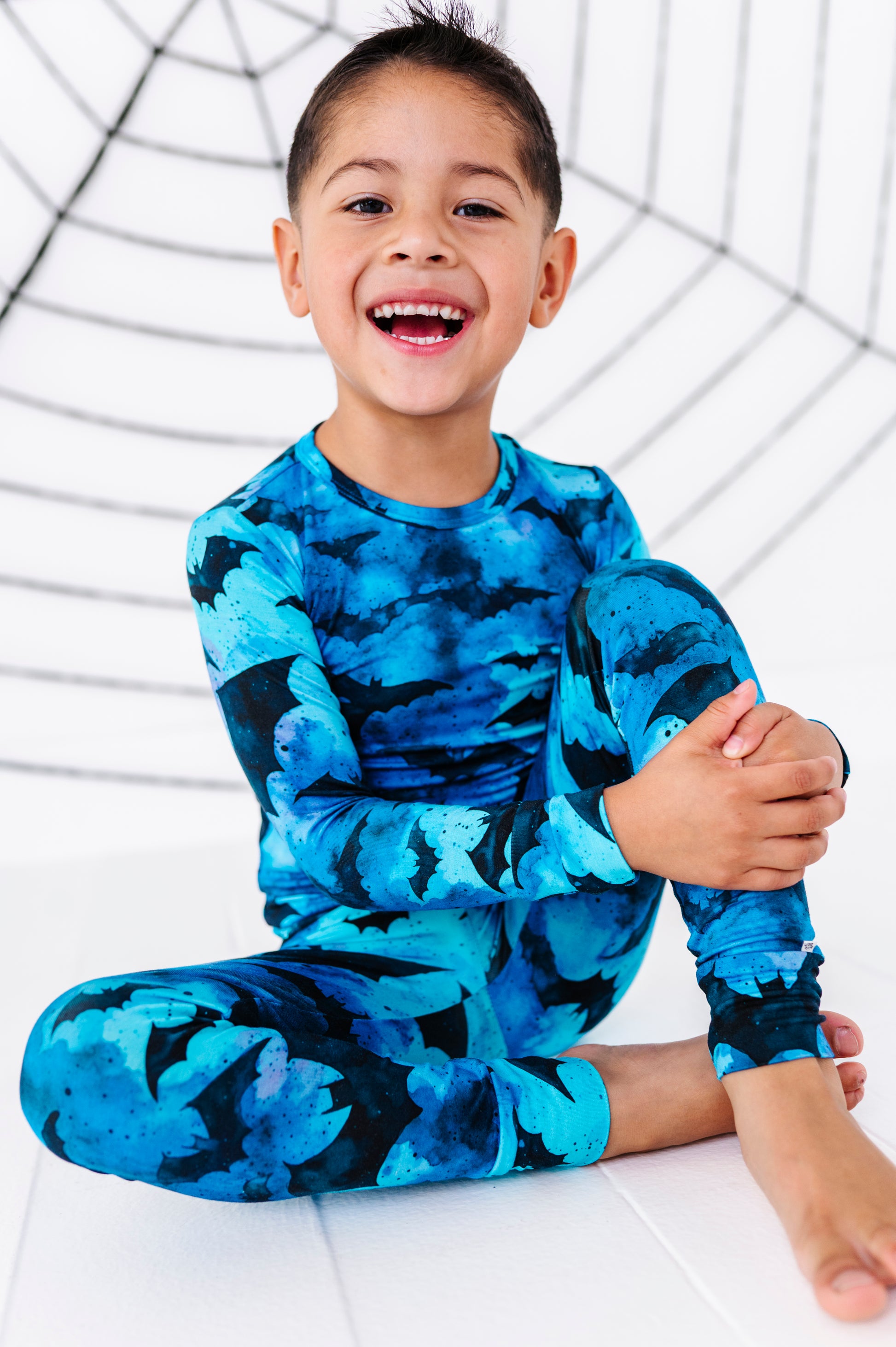 A young boy smiling and sitting cross-legged on a white floor, wearing a long-sleeve matching blue and black patterned shirt and pants set. A black spiderweb graphic is visible in the background.