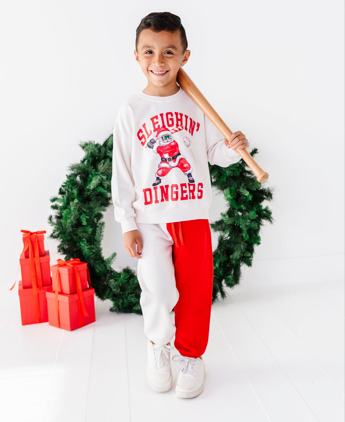 A young boy stands in a studio setting wearing a white long-sleeve sweatshirt with a red Santa graphic and the words 'SLEIGHIN' DINGERS', paired with white sweatpants and bright red joggers. He holds a wooden baseball bat over his shoulder, with a green Christmas wreath and red gift boxes behind him on a white background.
