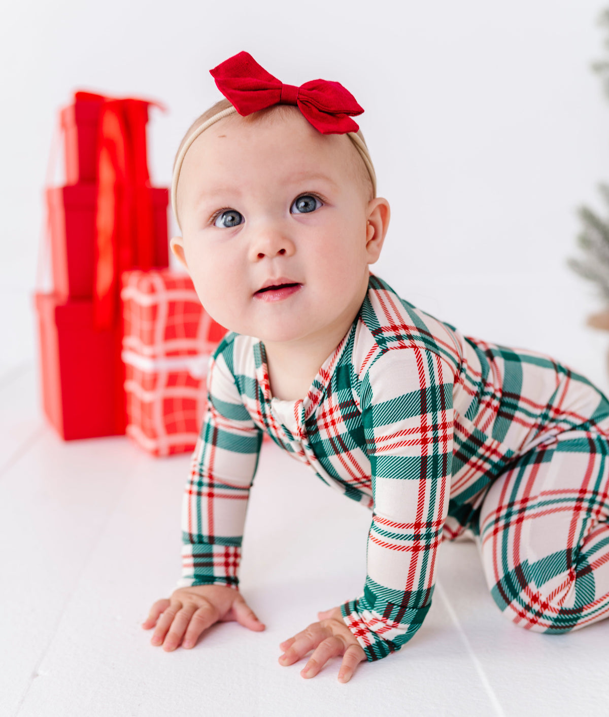 A baby crawling on a white floor wearing a green, red, and white plaid long-sleeve bamboo romper and a red bow headband, with red gift boxes in the background.