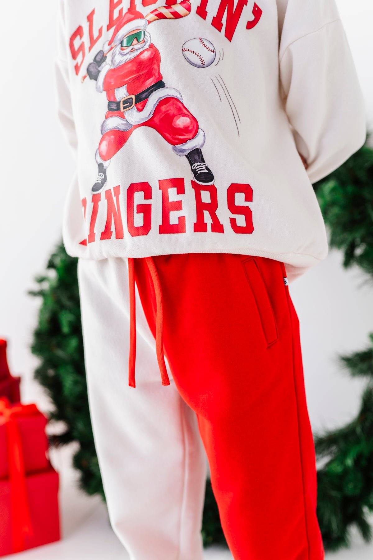 A person wearing a white sweatshirt with a red graphic of a baseball player dressed as Santa and the text 'SLEEPING SINGERS,' paired with bright red sweatpants. The photo is cropped from the chest down, with blurred greenery and red gift boxes in the background.