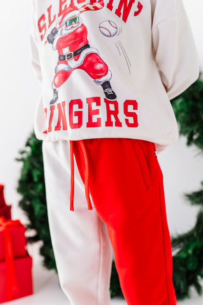 A person wearing a white sweatshirt with a red graphic of a baseball player dressed as Santa and the text 'SLEEPING SINGERS,' paired with bright red sweatpants. The photo is cropped from the chest down, with blurred greenery and red gift boxes in the background.