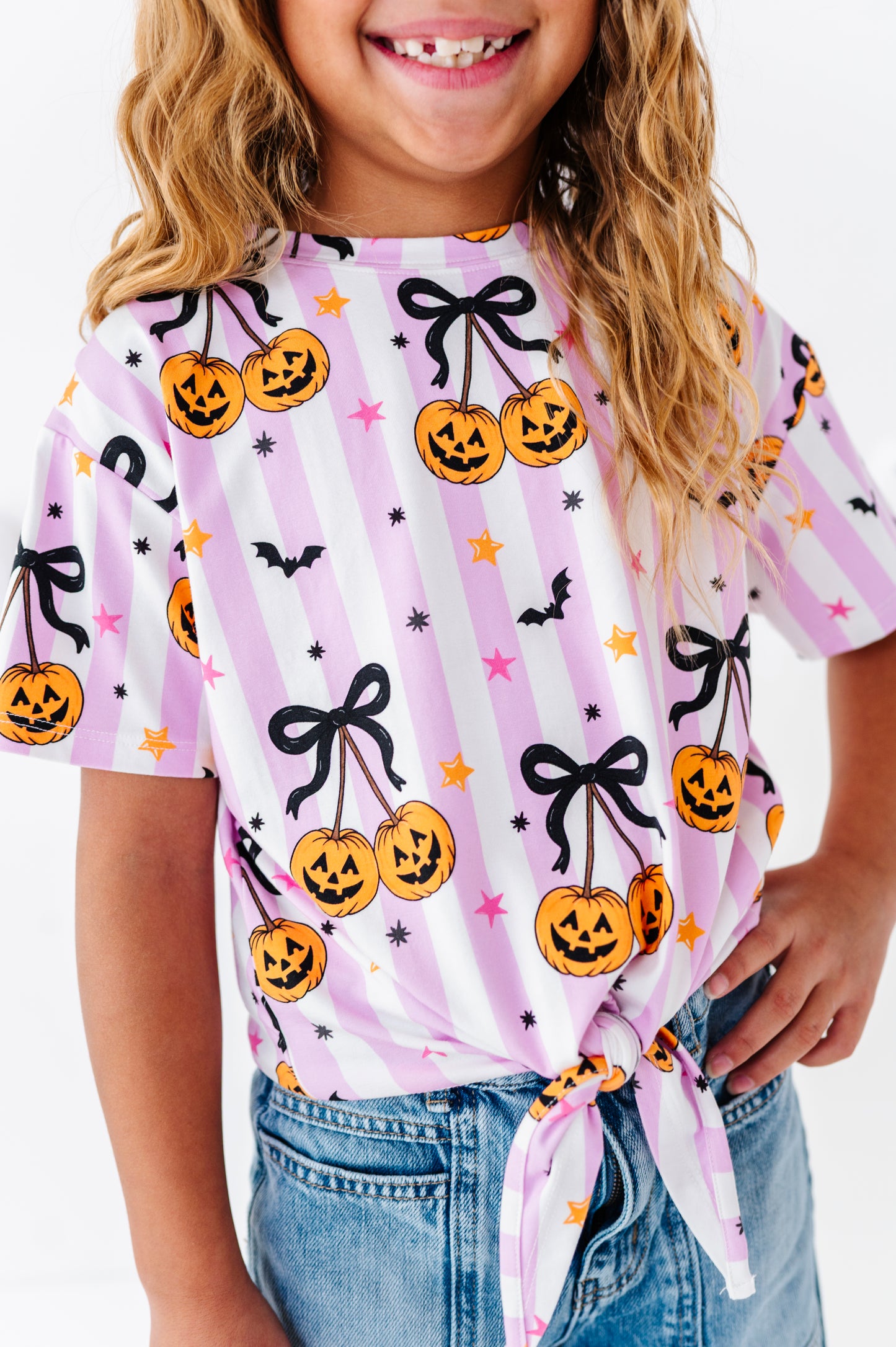 A child wearing a short-sleeve t-shirt with a pink and white striped Halloween pattern featuring jack-o'-lanterns, black bows, stars, and bats, paired with blue jeans.