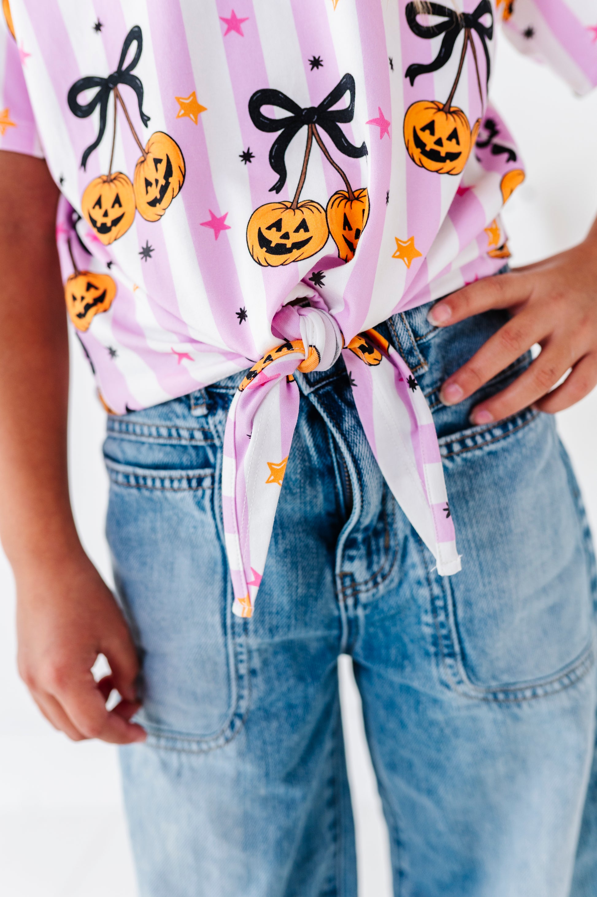 A close-up of a person wearing a pink and white striped t-shirt with a Halloween pumpkin print, tied at the waist with blue jeans.