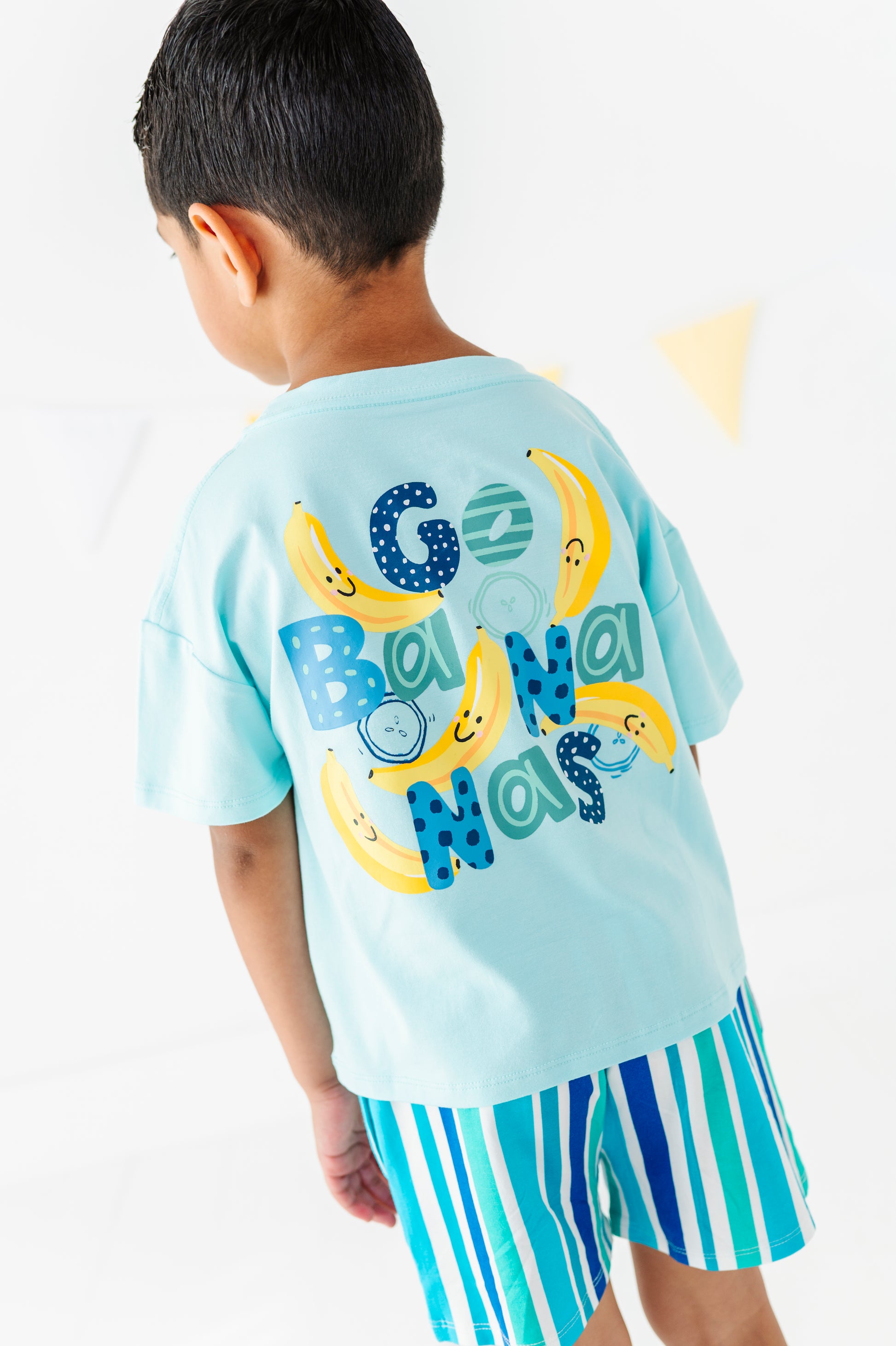 A child wearing a light blue t-shirt with a colorful 'Go Banana Man' graphic on the back and blue, green, and white striped shorts, shown from behind against a white background.