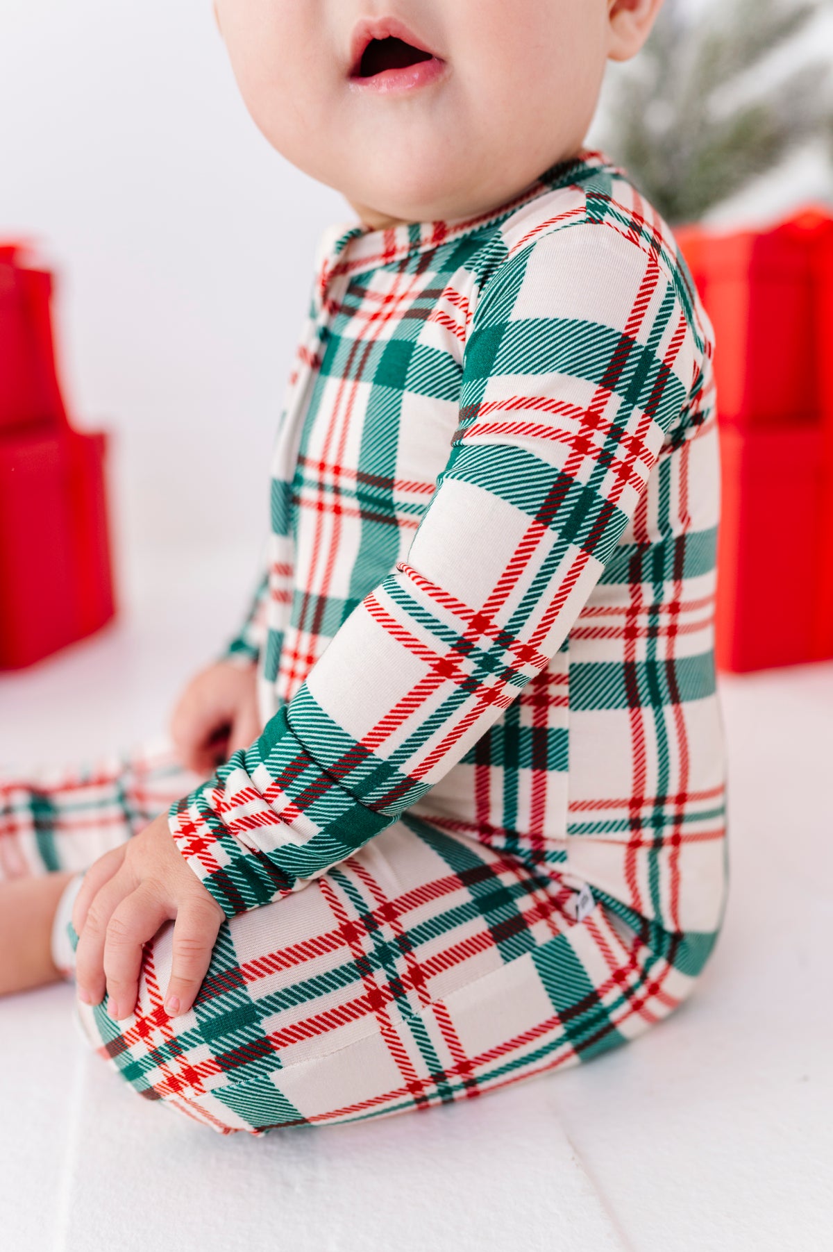 A baby wearing a long-sleeve holiday plaid romper in green, red, and white, sitting on a white surface with blurred red gift boxes in the background.