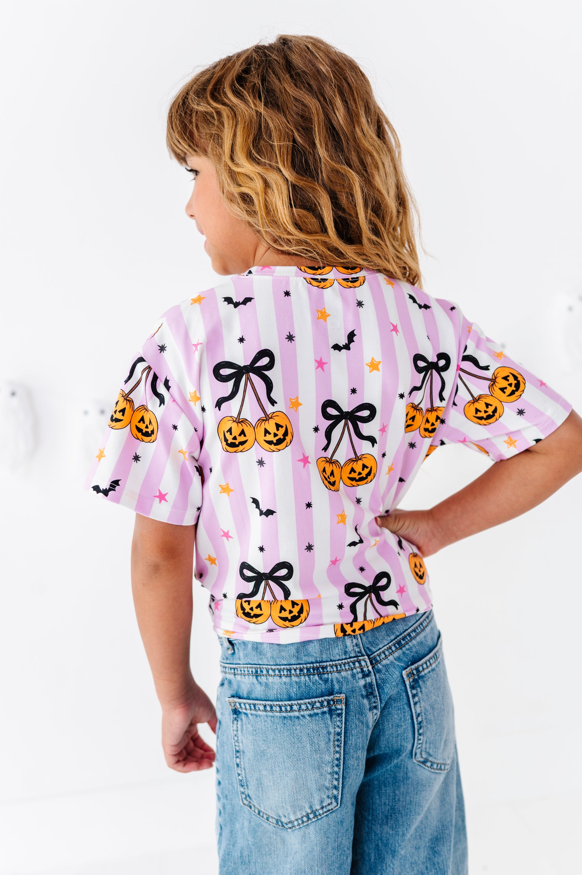 A child wearing a light pink striped short-sleeve t-shirt with a Halloween print of jack-o'-lanterns, black bows, and bats, paired with blue jeans, shown from the back against a white wall.
