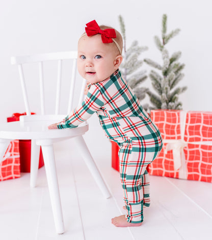 A baby standing on a white wooden floor next to a white chair, wearing a green, red, and white plaid holiday pajama set and a red bow headband. Christmas wrapped gifts and a small tree are in the background.