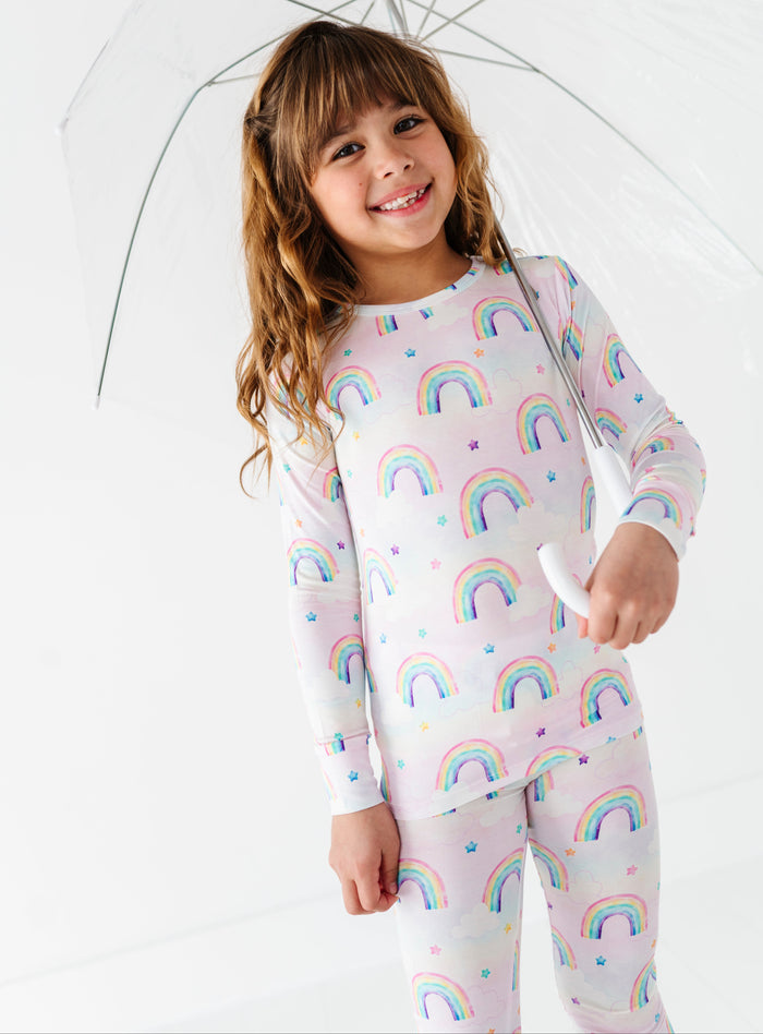 Child wearing a rainbow-patterned bamboo two-piece pajama outfit holding an umbrella on a white background