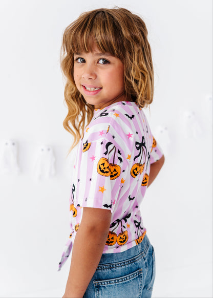 A young girl with wavy light brown hair looks back over her shoulder while wearing a pink and white striped short-sleeve t-shirt with a Halloween pumpkin print, paired with blue jeans against a white wall.