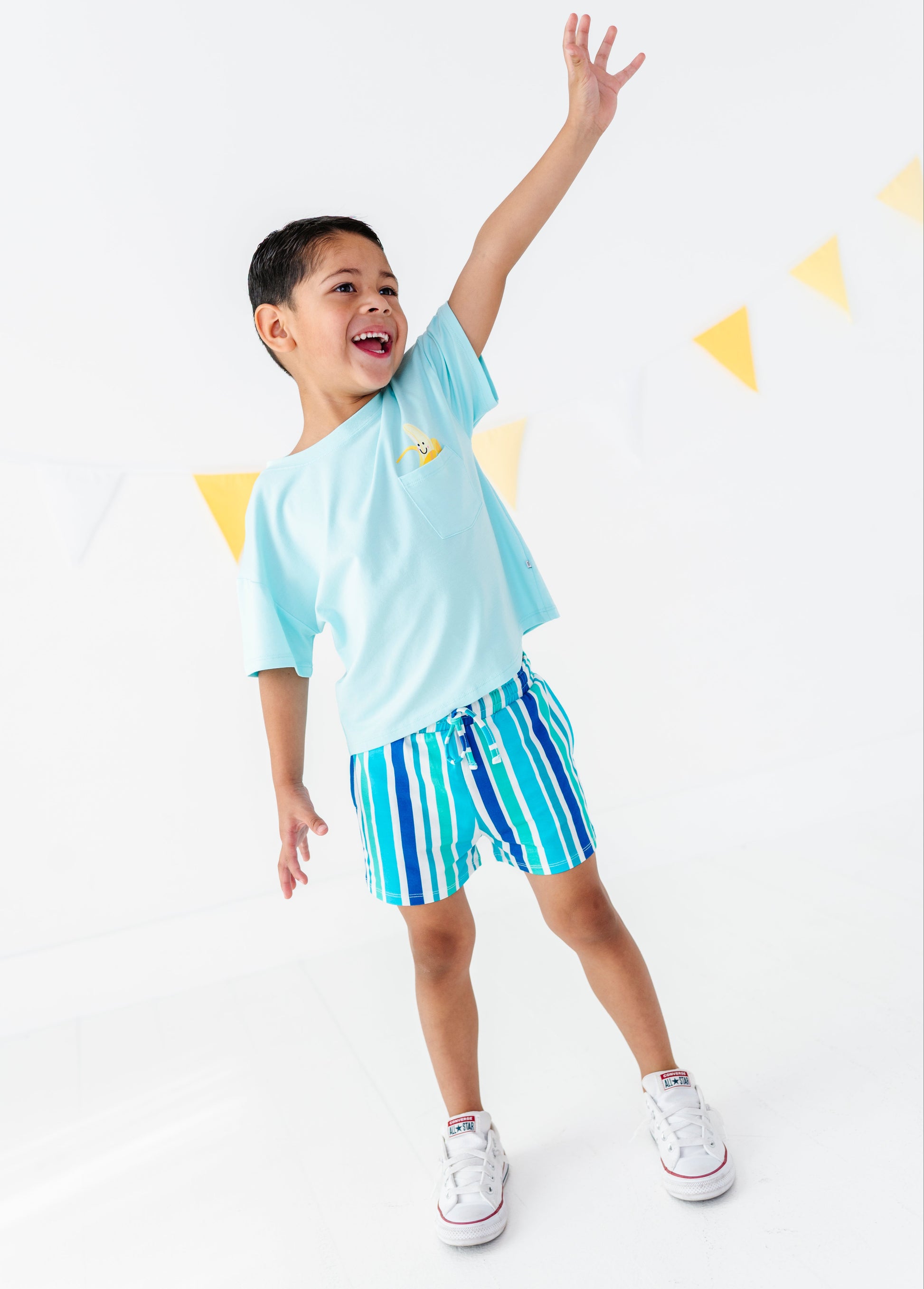 A young boy wearing a light blue t-shirt and blue and green striped swim trunks, posing with one arm raised against a white background with yellow bunting.