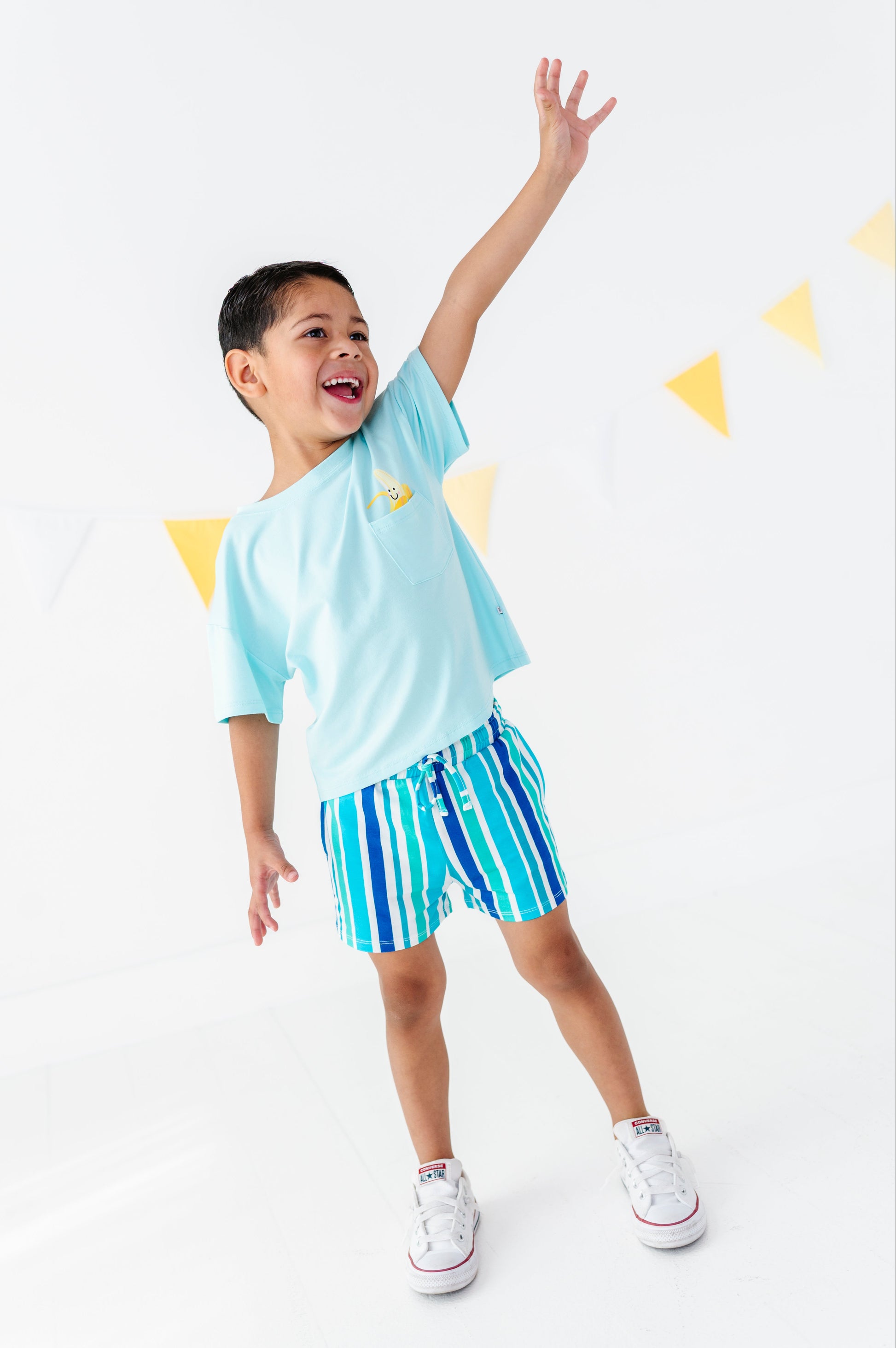 A young boy wearing a light blue t-shirt and blue and green striped swim trunks, posing with one arm raised against a white background with yellow bunting.