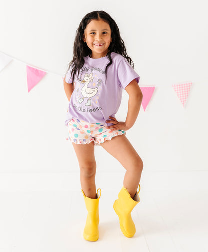Child wearing a purple shirt with a goose design, polka dot shorts, and yellow rain boots on a white background.