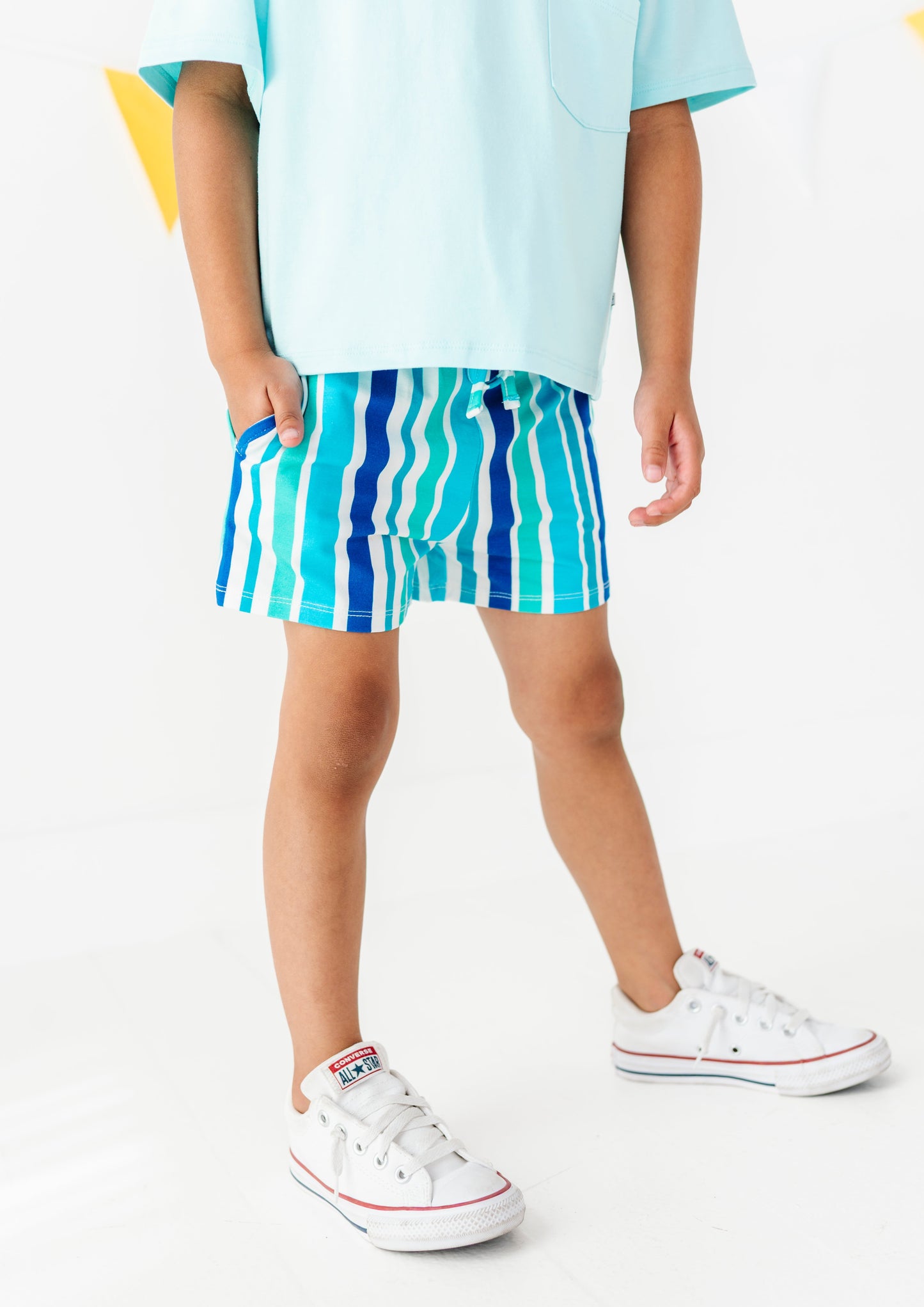 A child wearing blue and turquoise striped swim trunks, a light blue t-shirt, and white low-top sneakers, standing against a white studio background.