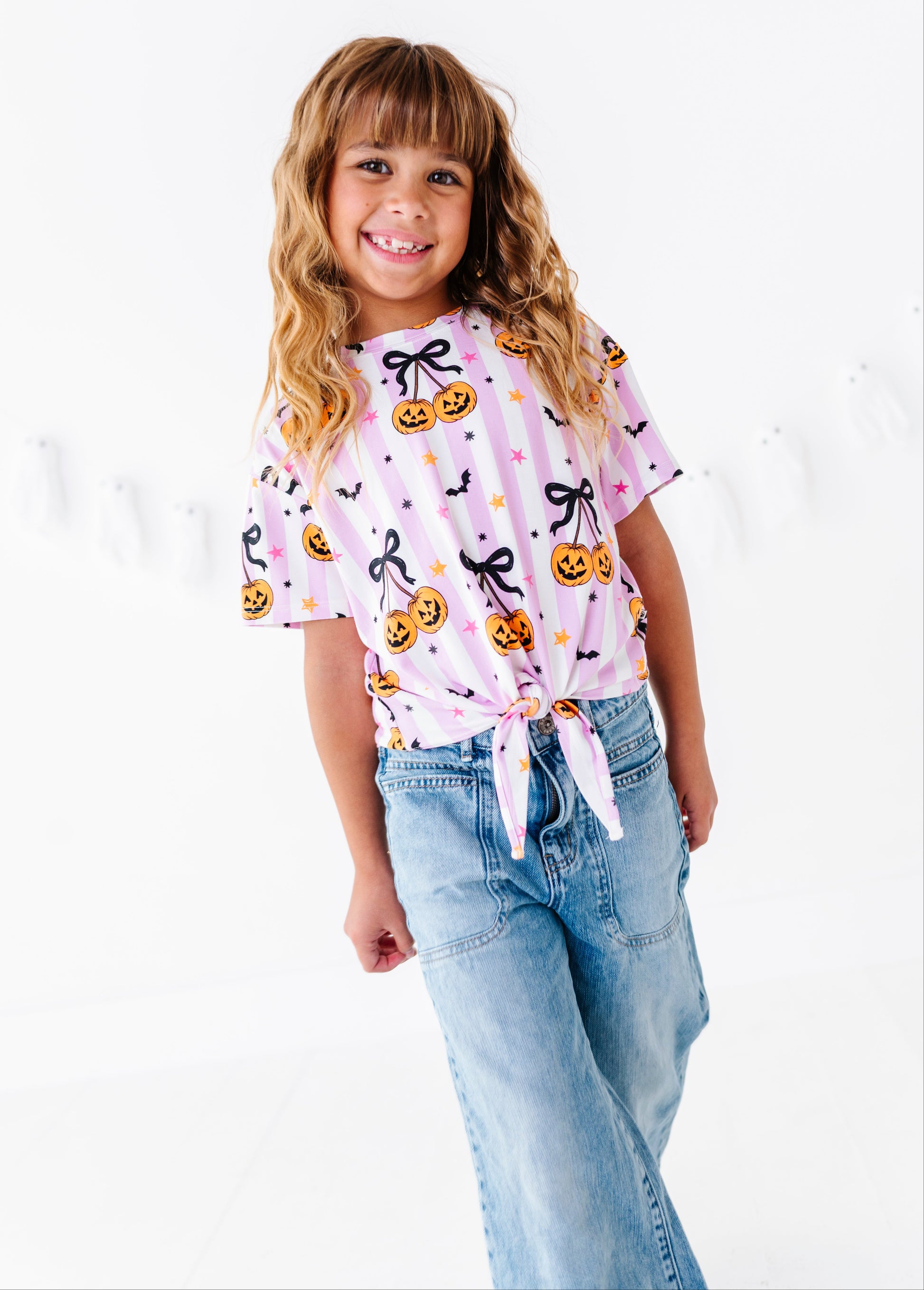 A young girl with blonde hair smiling and wearing a pink short-sleeve tie-front t-shirt with a Halloween pumpkin print, paired with light-wash blue jeans, against a white studio background.
