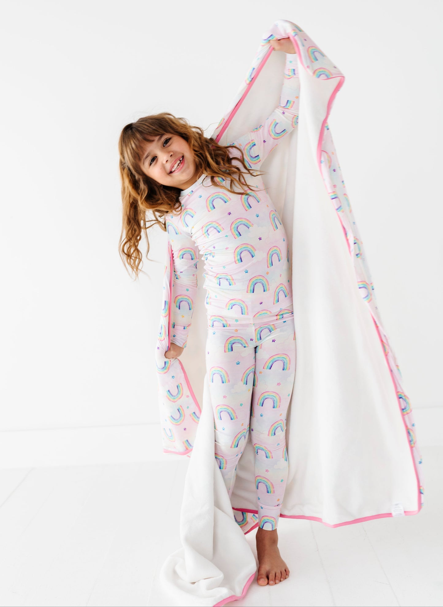 Young girl holding a large bamboo plush blanket with pink trim and watercolor rainbow patterns on a white background
