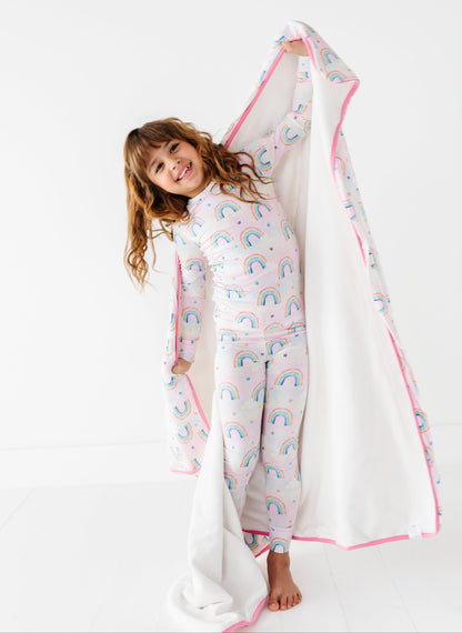 Young girl holding a large bamboo plush blanket with pink trim and watercolor rainbow patterns on a white background