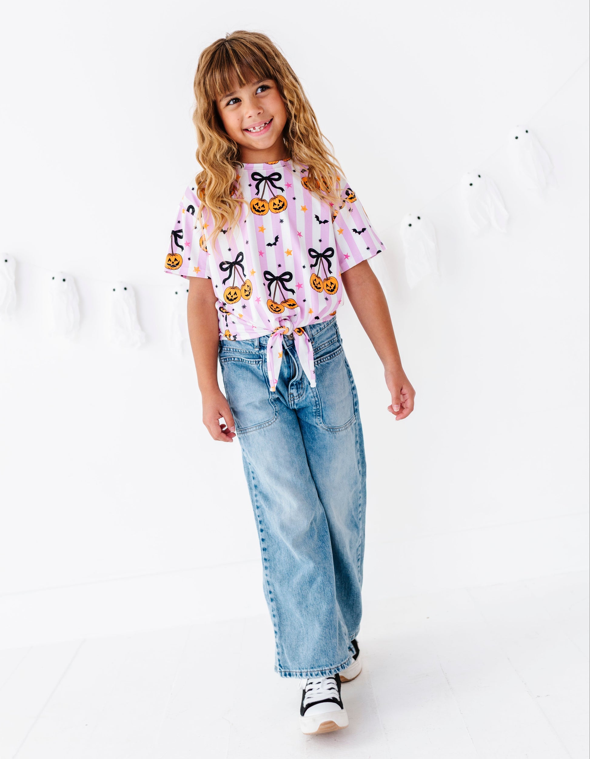 A young girl with blonde hair is smiling and posing in a white studio setting, wearing a light pink short-sleeve t-shirt with a pumpkin and bow pattern, light-wash wide-leg jeans, and white sneakers.