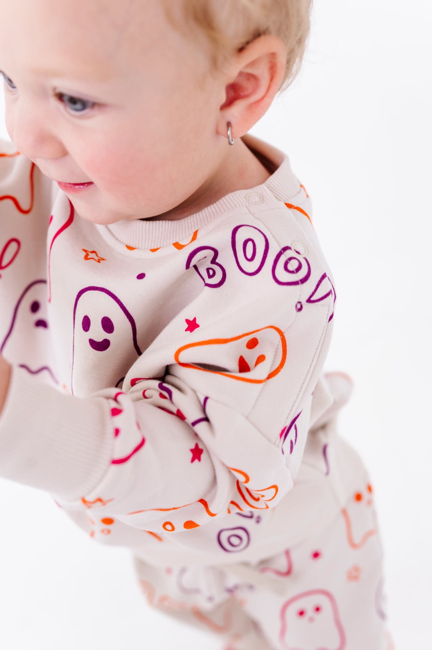 A close-up of a baby wearing a cream-colored long-sleeve sweatshirt with a colorful Halloween-themed print of ghosts, stars, and the word 'BOO'.