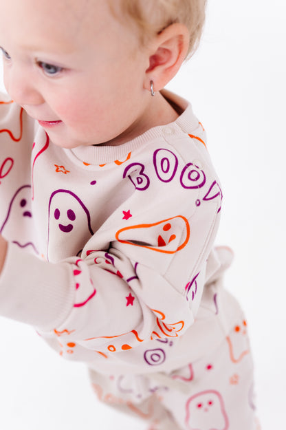 A close-up of a baby wearing a cream-colored long-sleeve sweatshirt with a colorful Halloween-themed print of ghosts, stars, and the word 'BOO'.