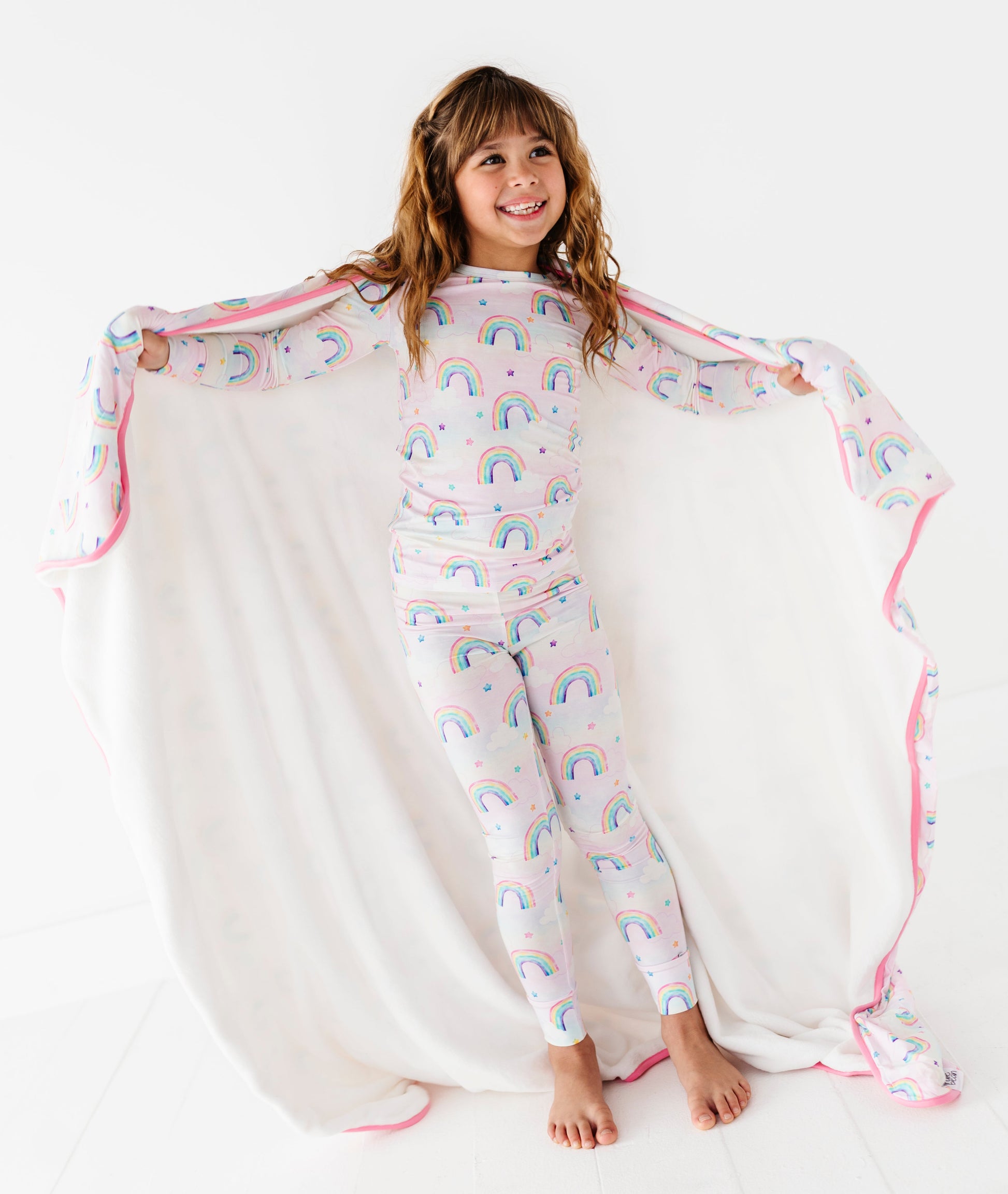 Child wearing a rainbow-patterned bamboo pajama set with a matching bamboo blanket draped over them on a white background
