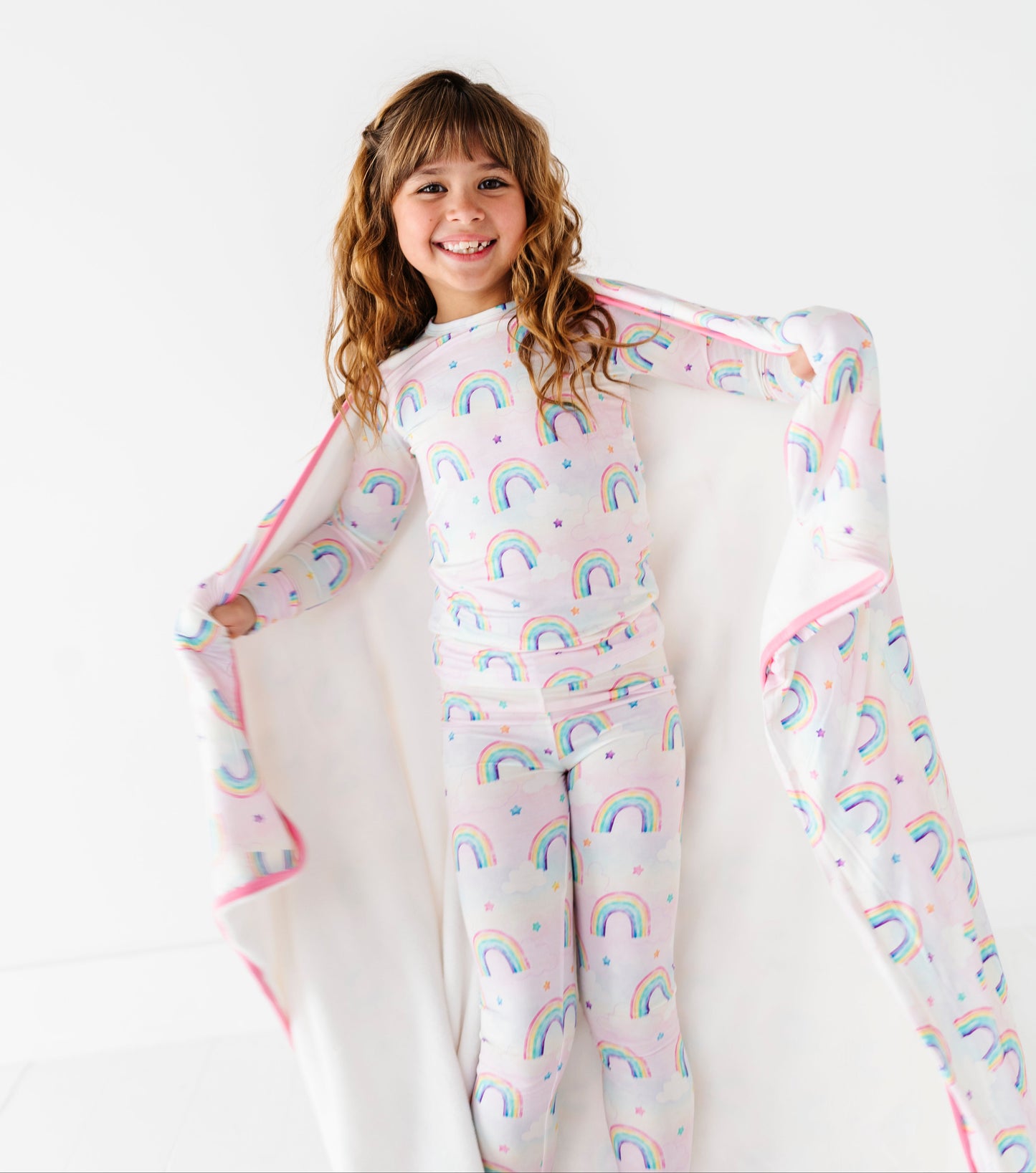 Child wearing a rainbow-patterned bamboo pajama set with a matching bamboo blanket on a white background