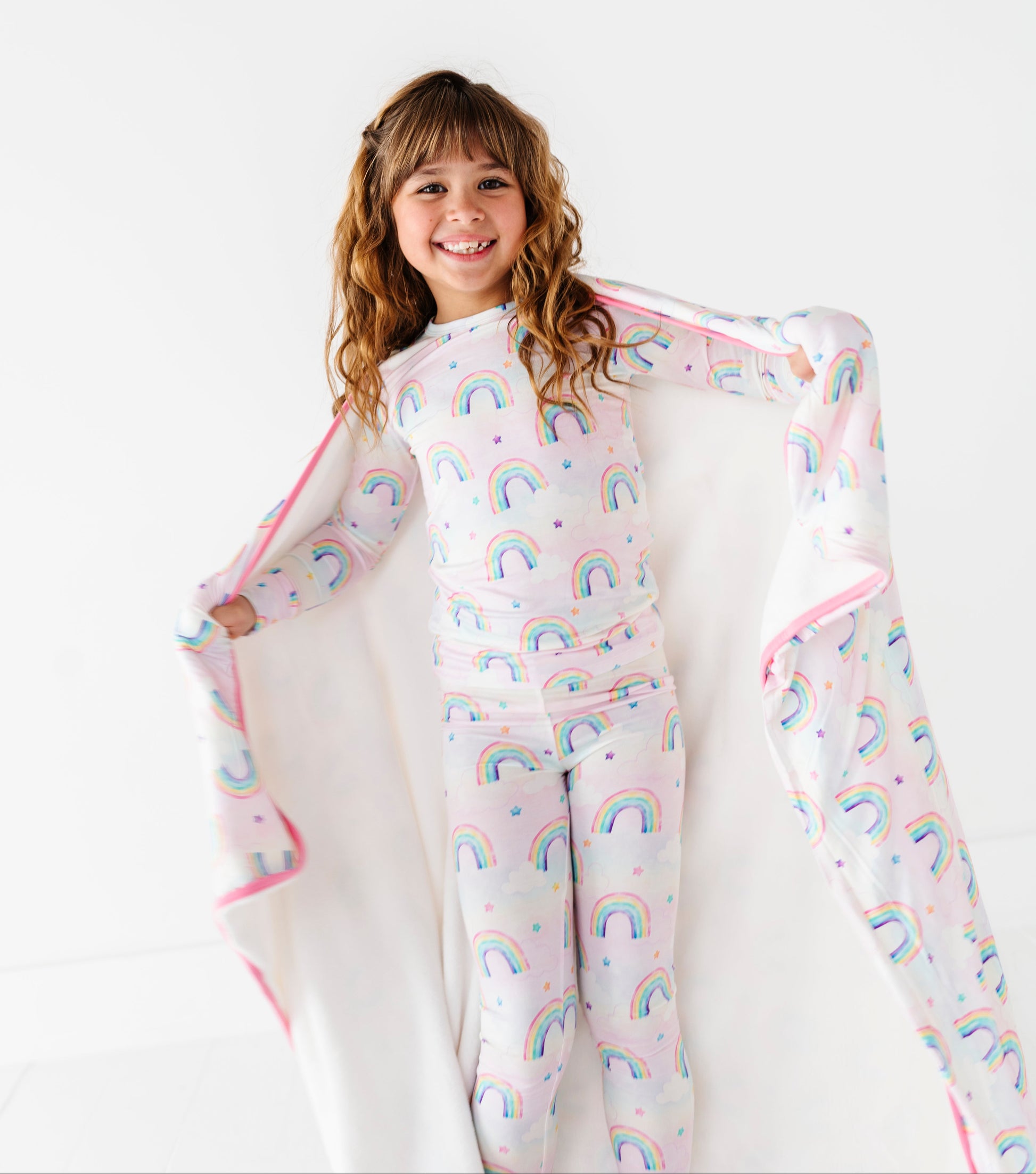 Child wearing a rainbow-patterned bamboo pajama set with a matching bamboo blanket on a white background