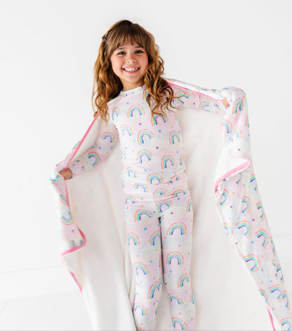 Child wearing a rainbow-patterned bamboo pajama set with a matching bamboo blanket on a white background