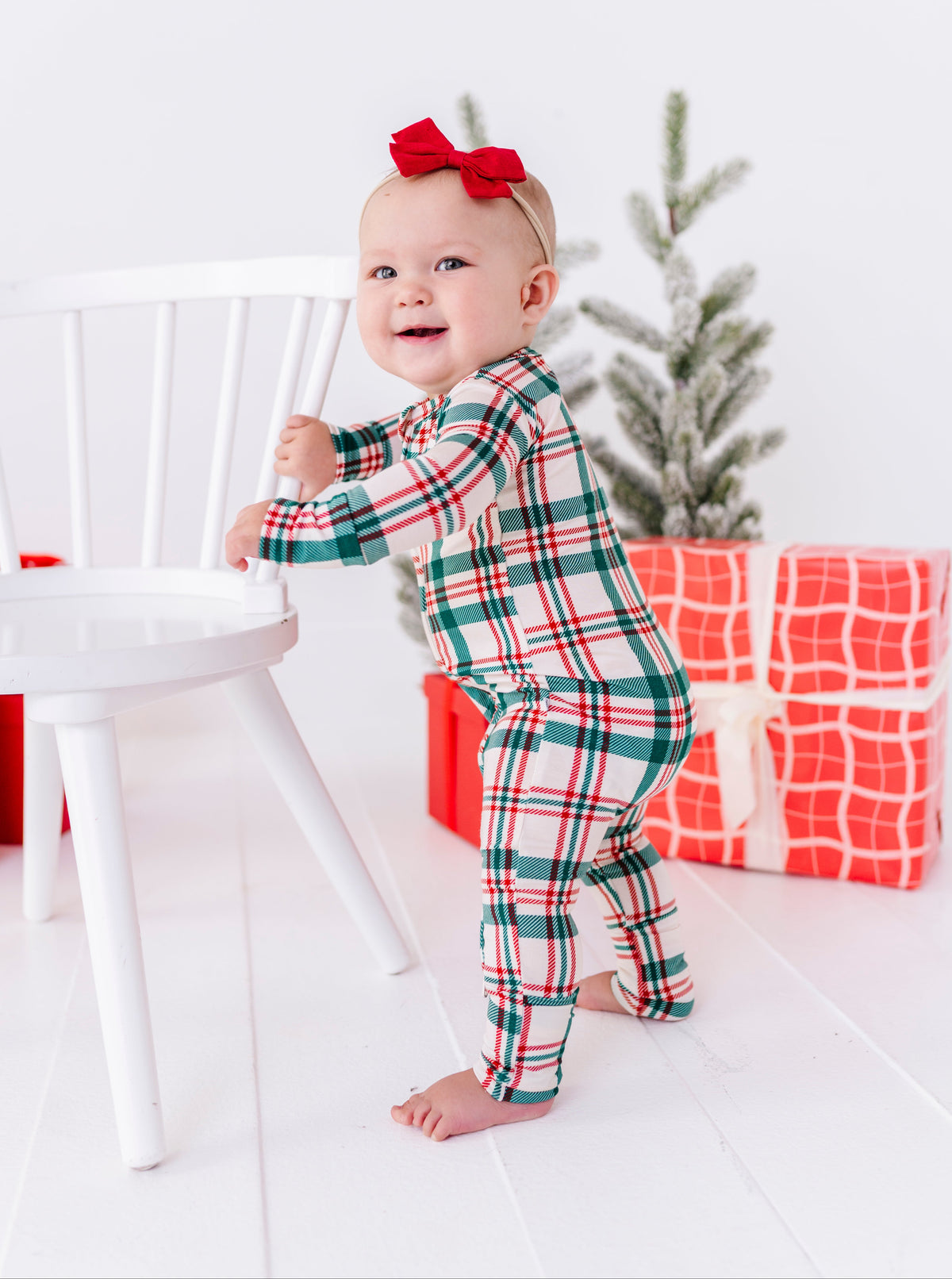 A baby wearing a festive green, red, and white plaid pajama set and a red bow headband stands on a white floor next to a white chair, with wrapped Christmas presents and a small tree in the background.