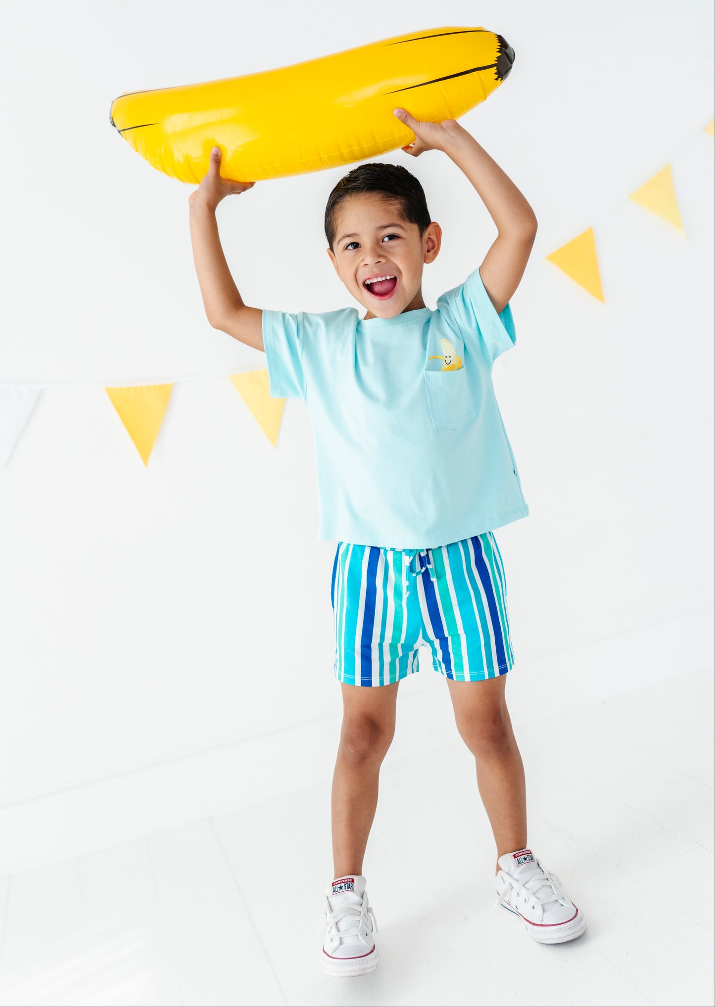 A young boy wearing a light blue t-shirt and blue and white striped swim trunks stands on a white background, holding a yellow inflatable pool float above his head.