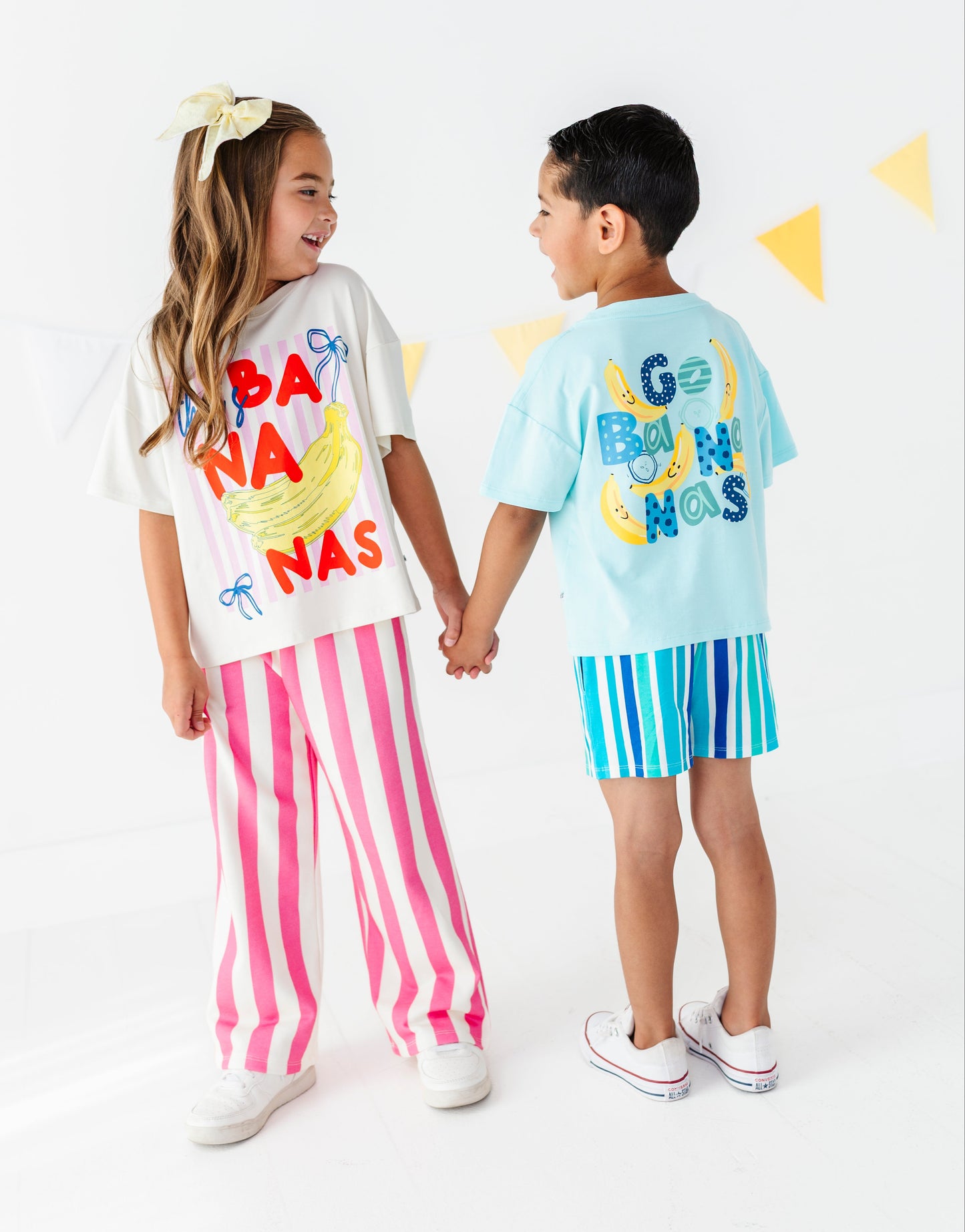 Two children holding hands, wearing matching tropical banana-themed outfits. The girl wears a white graphic t-shirt with 'BANANAS' in red and yellow, paired with pink and white vertical stripes.