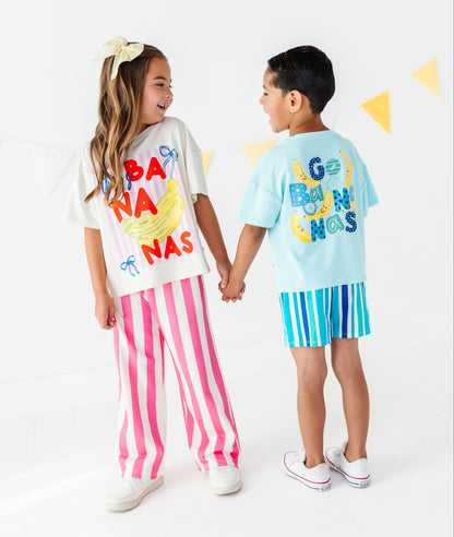 Two children holding hands, wearing matching tropical banana-themed outfits. The girl wears a white graphic t-shirt with 'BANANAS' in red and yellow, paired with pink and white vertical stripes.