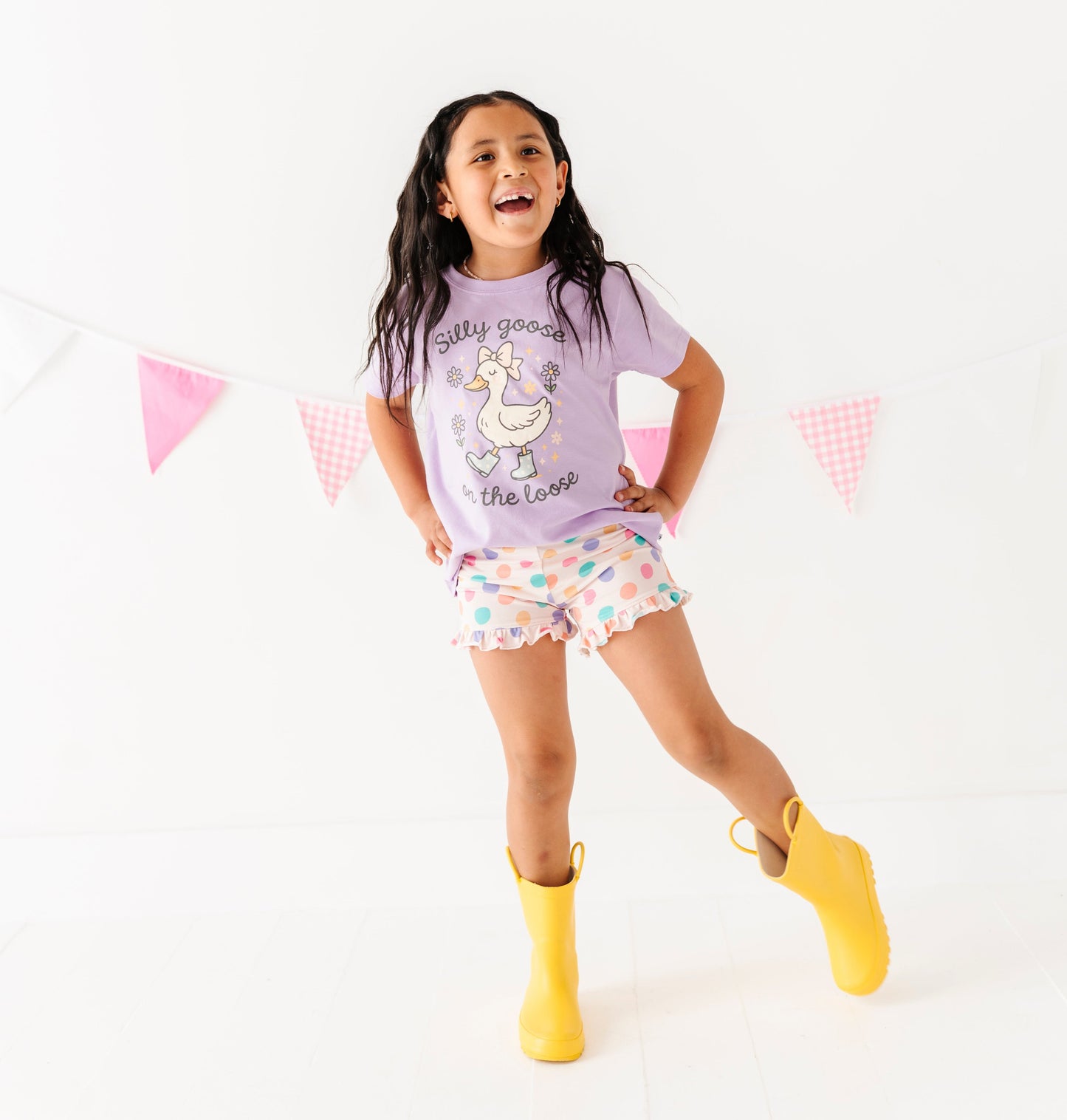 Child wearing a purple t-shirt with a unicorn design, patterned bamboo shorts, and yellow boots on a white background.
