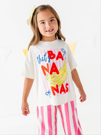A young girl with a yellow bow in her hair is wearing a white graphic t-shirt that reads 'that's BANanas' in red, blue, and yellow, paired with pink and white striped pants, against a clean white background.