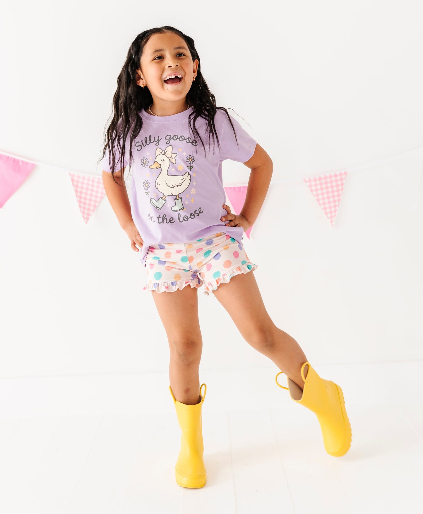 Child wearing a purple t-shirt with a goose design, polka dot shorts, and yellow rain boots on a white background.