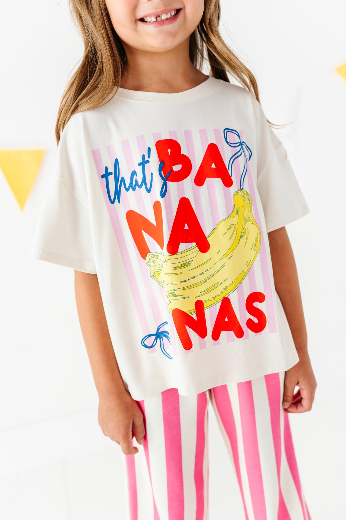 A young girl wearing a white graphic t-shirt with a colorful 'that's BANanas' print featuring a banana illustration, paired with pink and white striped pants.