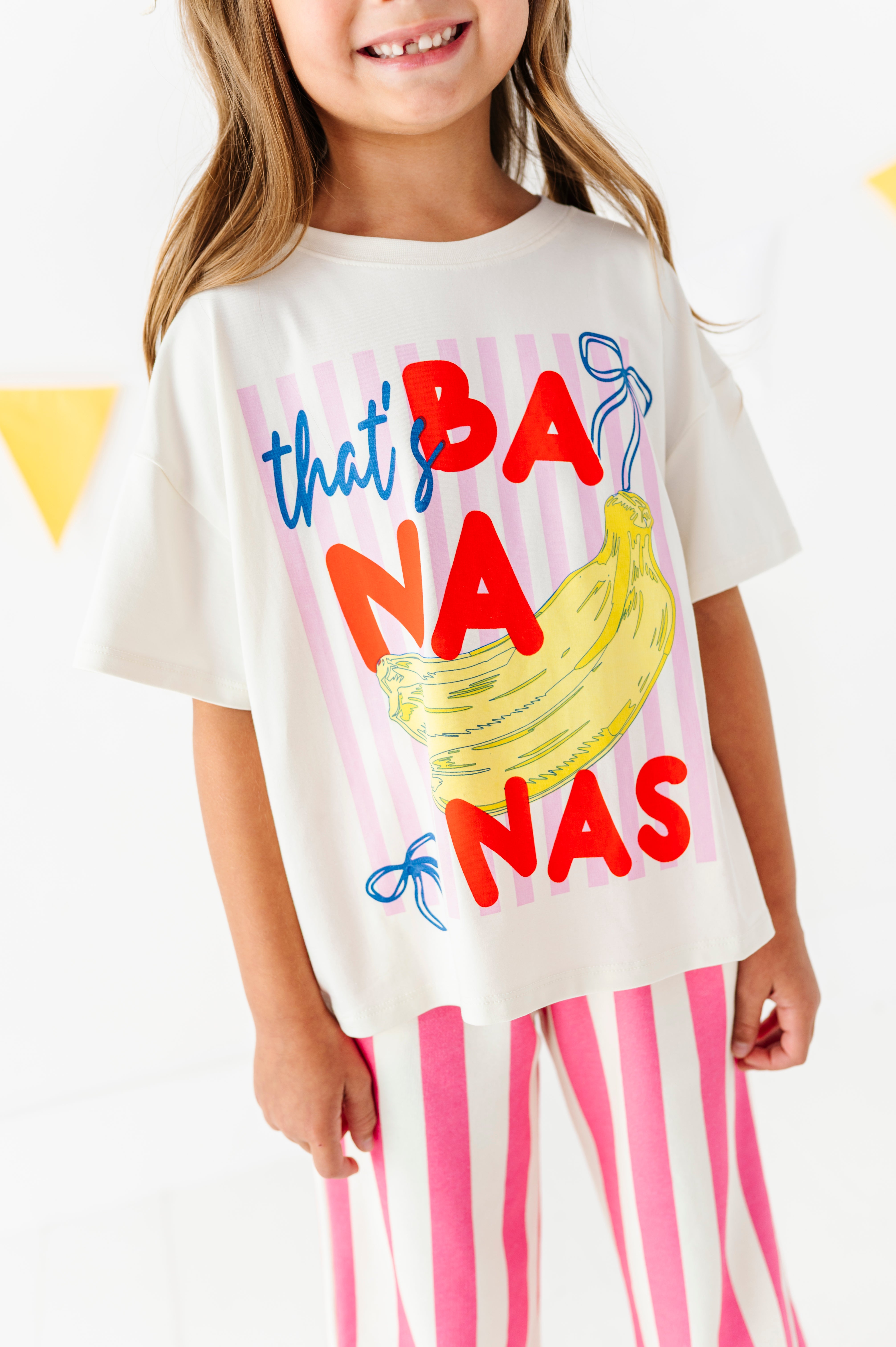 A young girl wearing a white graphic t-shirt with a colorful 'that's BANanas' print featuring a banana illustration, paired with pink and white striped pants.
