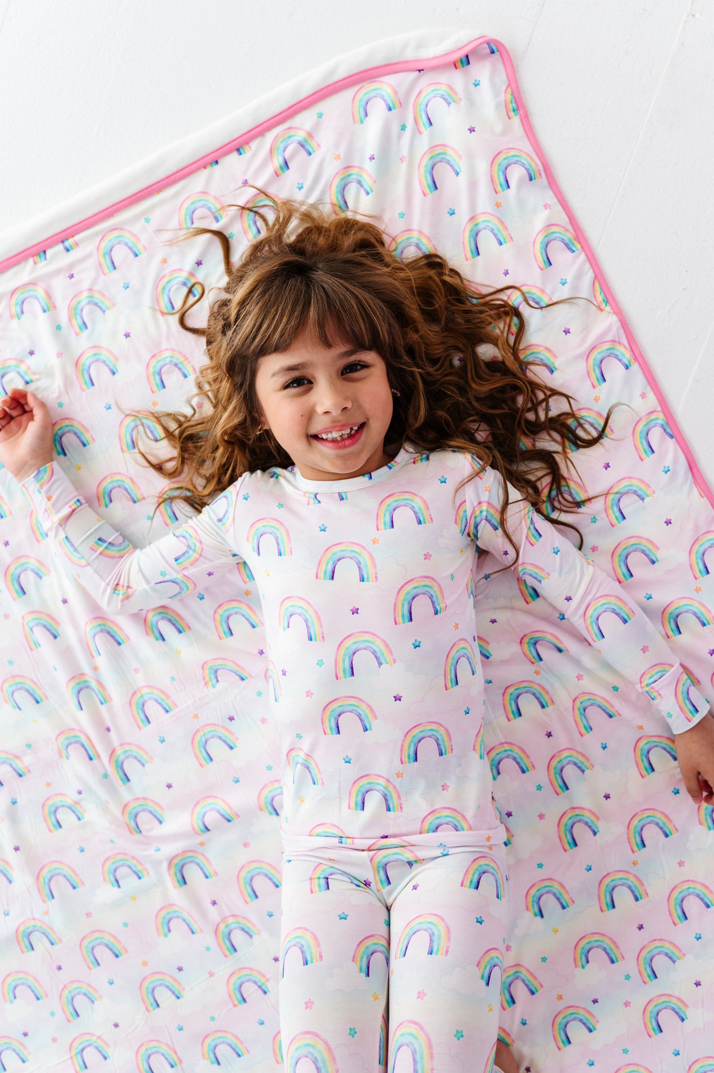 Child wearing a rainbow-patterned bamboo two-piece pajama set lying on a matching bamboo blanket.