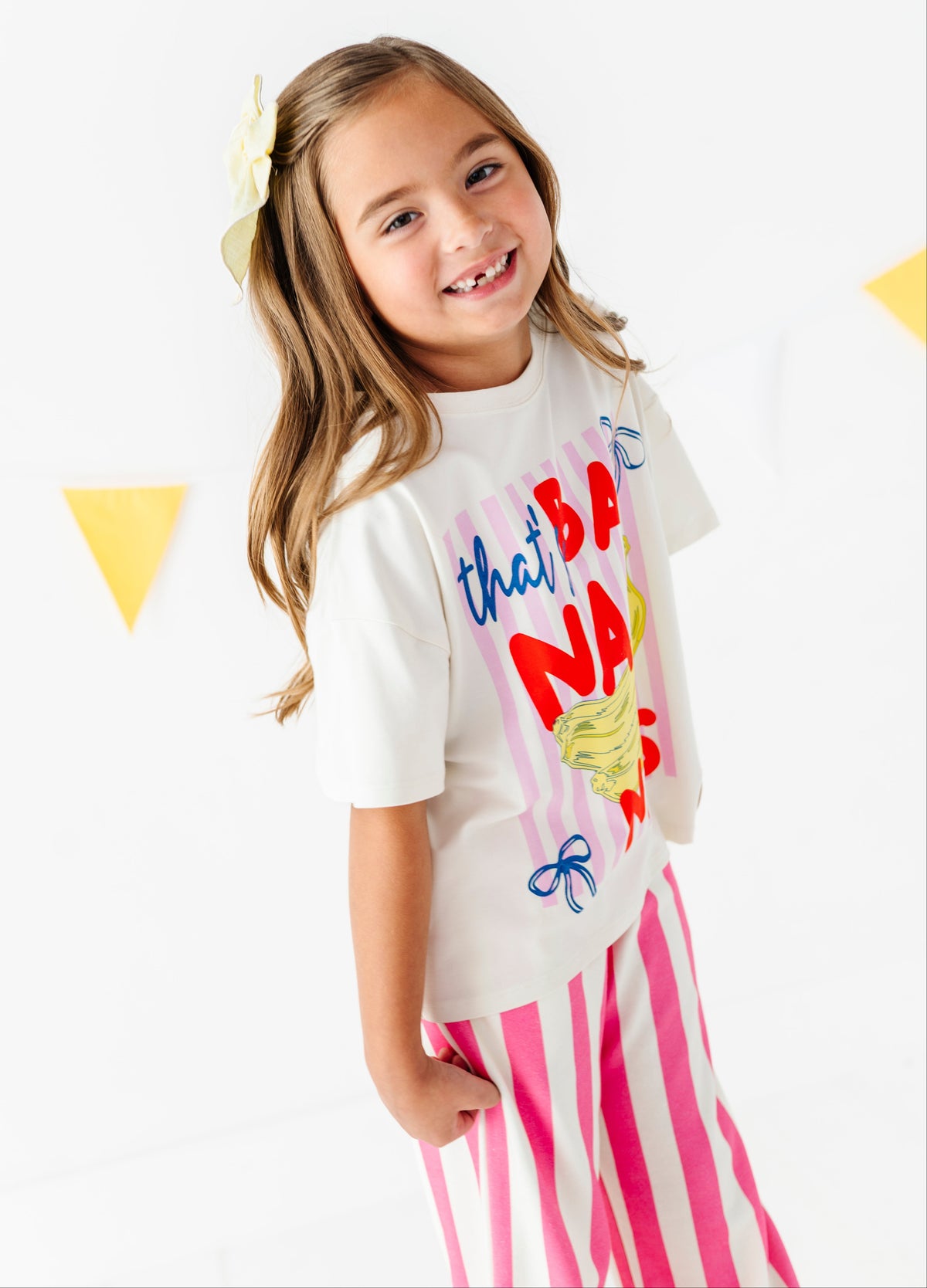 A young girl with long light brown hair wearing a white graphic t-shirt and pink and white striped pants, smiling and looking to the side against a white background with yellow bunting.