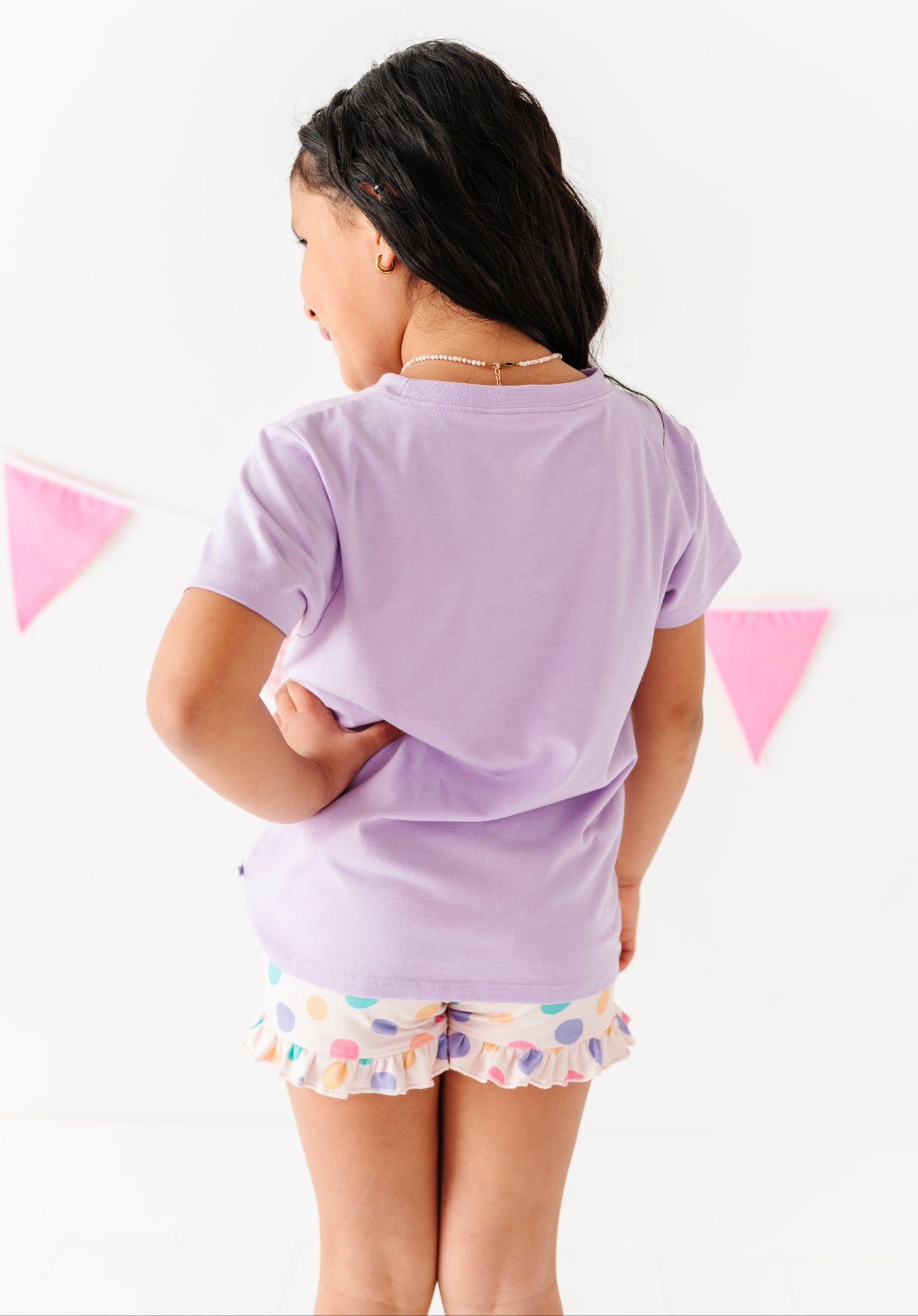 Girl wearing a light purple t-shirt and colorful shorts on a white background with pink flags.